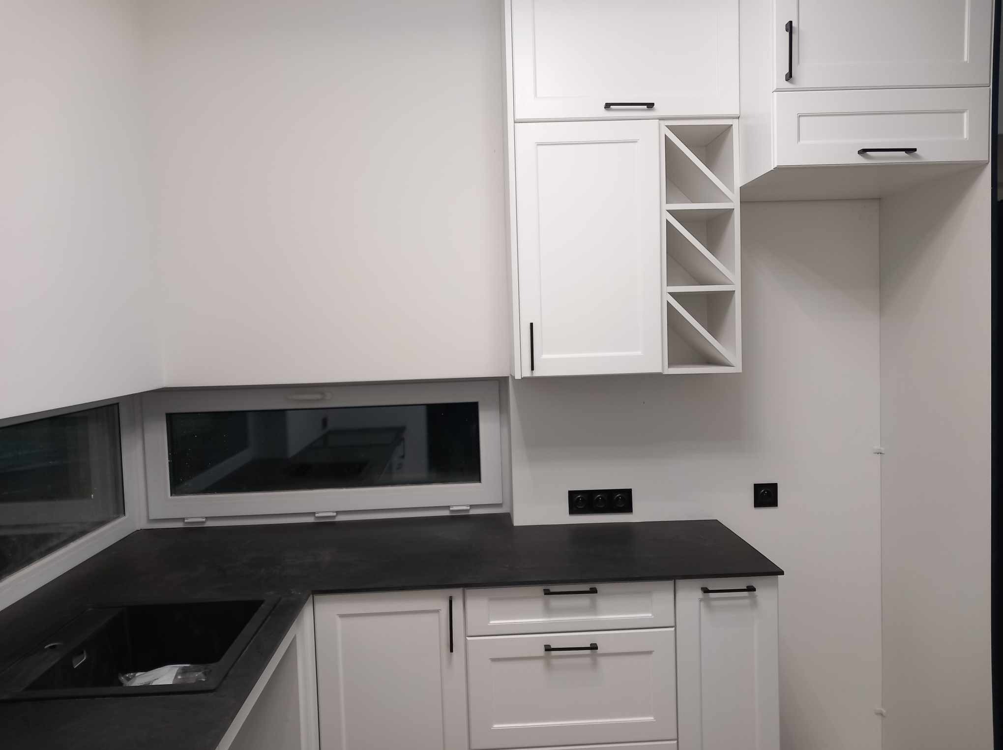 Nowoczesna, biała kuchnia z czarnym blatem i minimalistycznymi uchwytami, z oknem nad zlewem i półkami na wino.