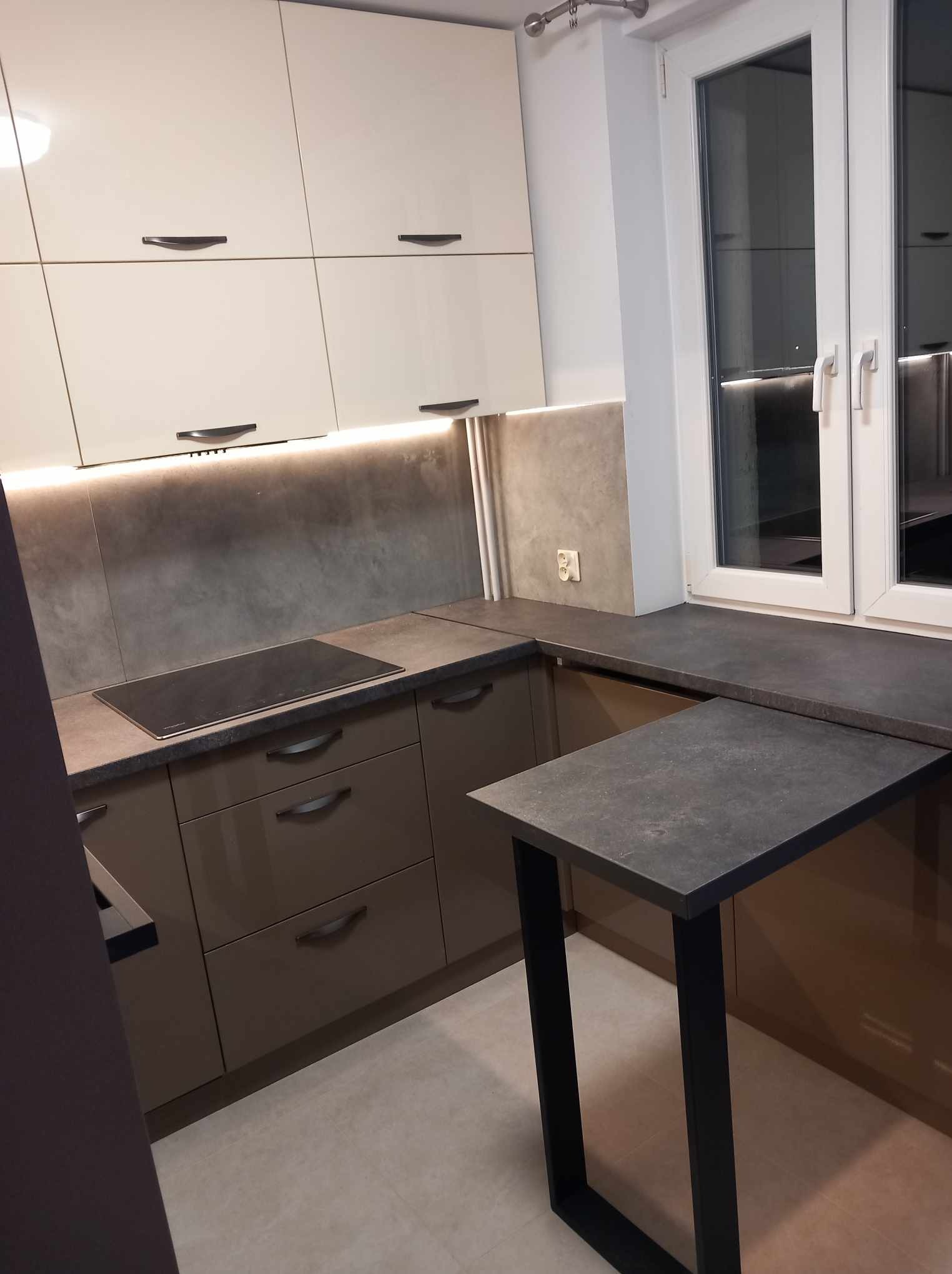 Nowoczesna kuchnia z beżowymi szafkami górnymi, brązowymi dolnymi, szarym blatem i wysuwanym, czarnym stołem, oświetlona listwą LED pod szafkami.