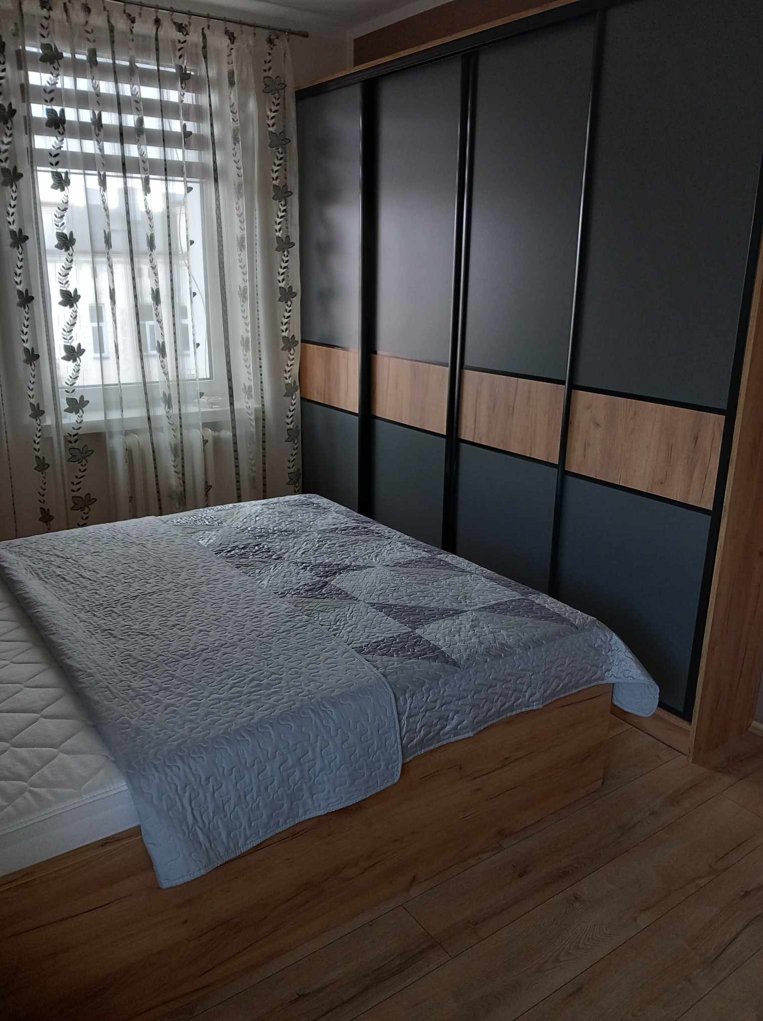 Sypialnia z łóżkiem i przesuwną szafą wnękową z czarnymi drzwiami i drewnianymi panelami, widok na okno z firankami w tle.