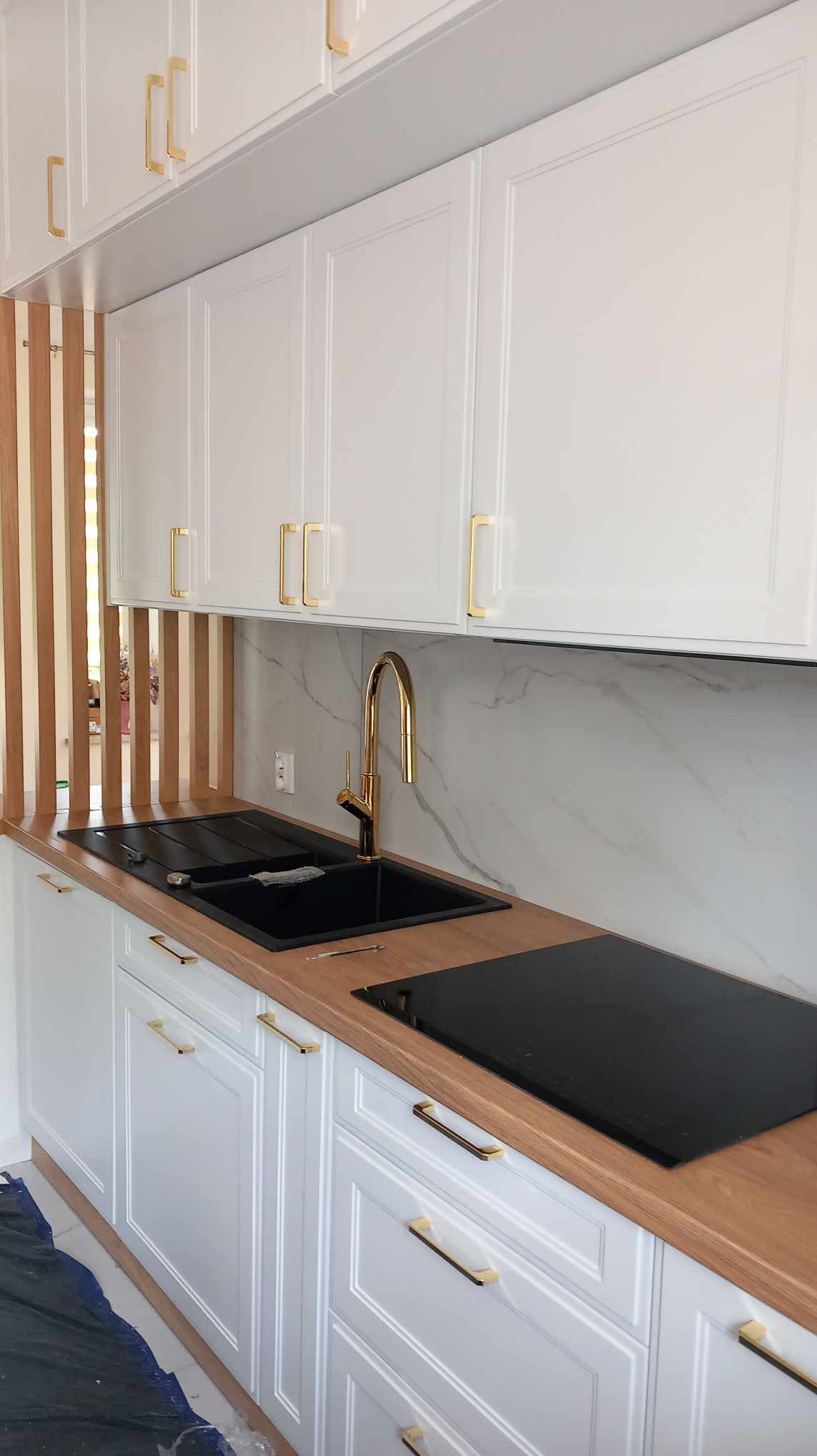 Białe szafki kuchenne z frezowanymi frontami i złotymi uchwytami, czarny zlew z baterią w kolorze złota, drewniany blat i płyta indukcyjna, marmurowy panel ścienny oraz drewniana ażurowa ścianka...
