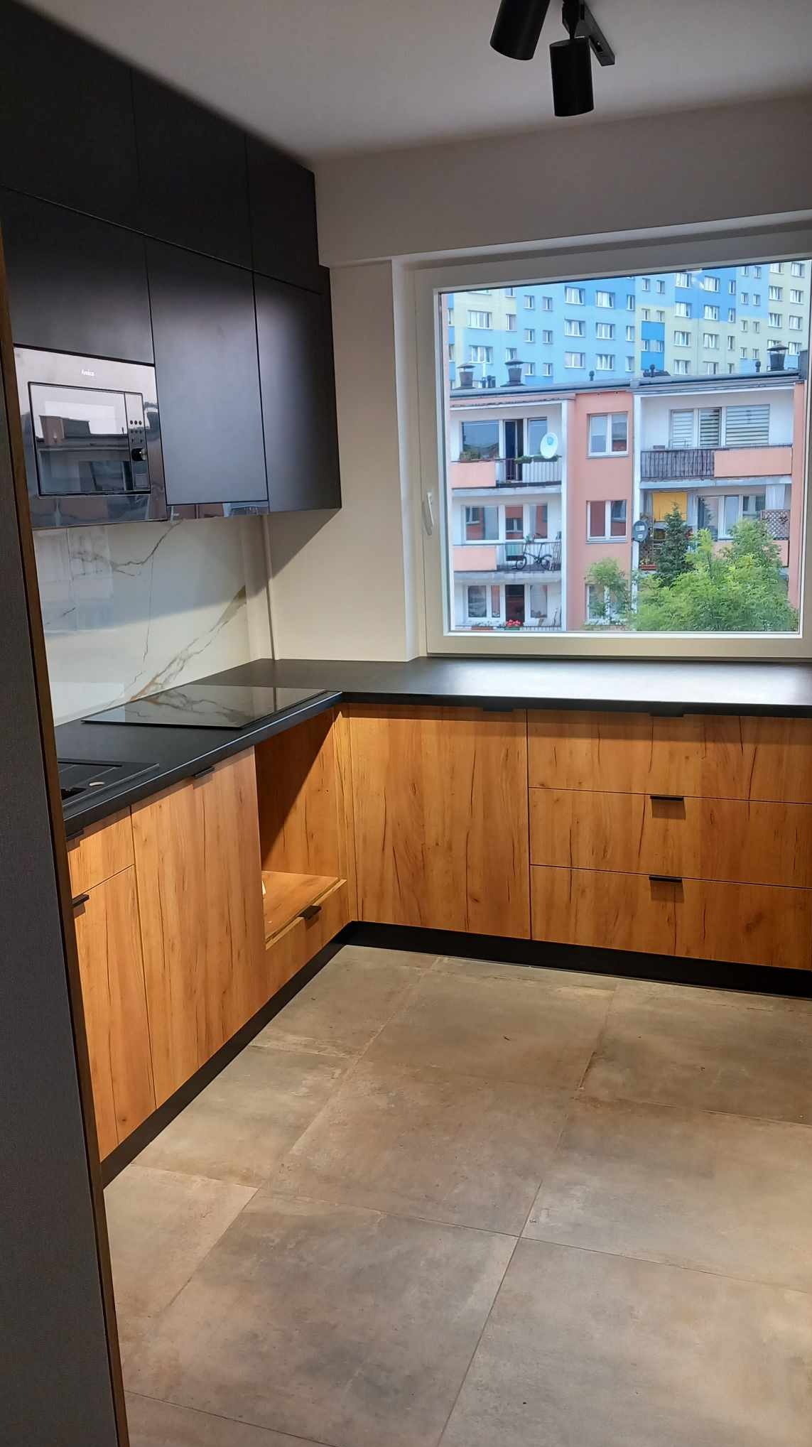 Nowoczesna kuchnia z drewnianymi szafkami dolnymi i czarnymi szafkami górnymi, czarnym blatem i oknem z widokiem na kolorowe bloki mieszkalne.