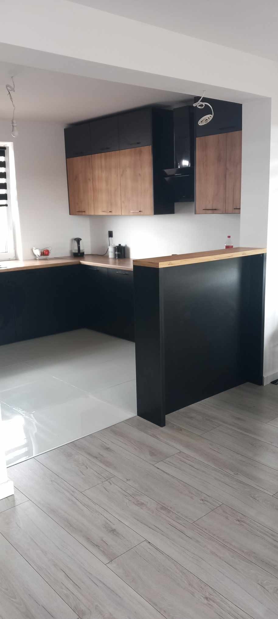 Nowoczesna kuchnia z czarnymi dolnymi szafkami, drewnianymi górnymi szafkami i blatem, oraz czarnym okapem. Białe ściany i podłoga z płytek imitujących drewno.