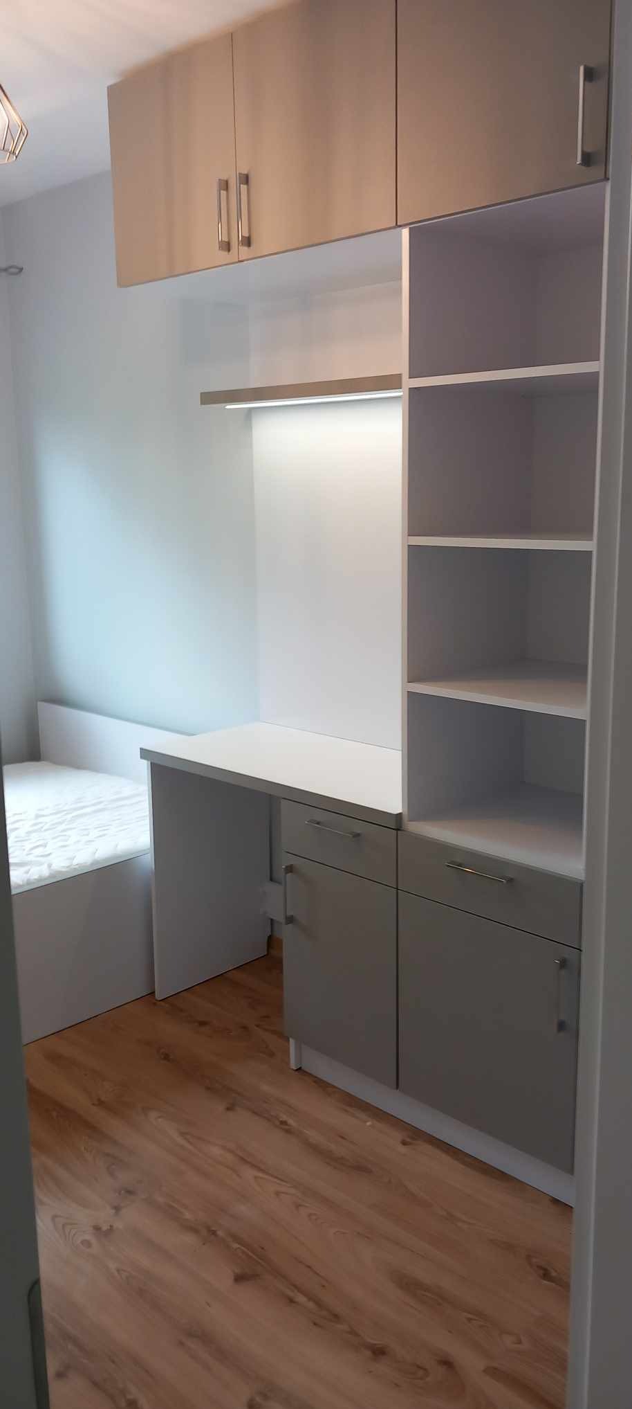Pokój z zabudową meblową: biurko z szufladami, półki i szafki górne w odcieniach bieli i szarości, dopasowane do łóżka, na podłodze panele.