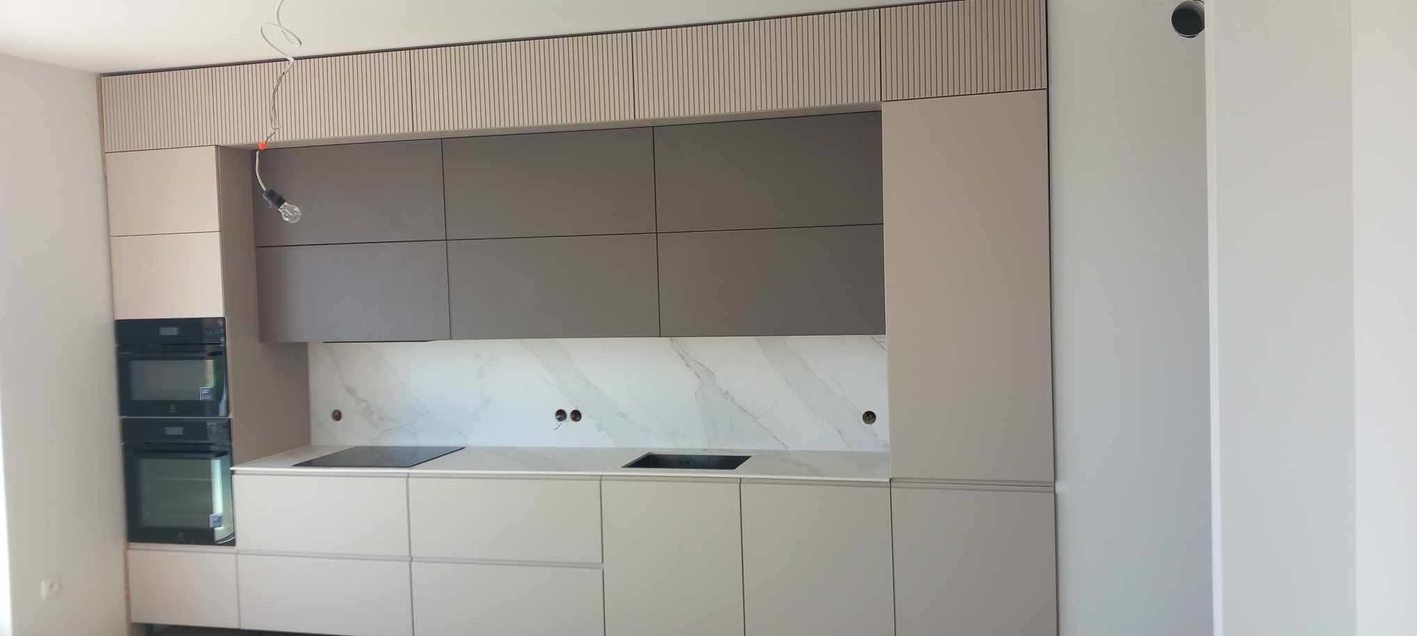 Nowoczesna kuchnia na wymiar z frontami w odcieniach szarości i beżu, marmurowy backsplash, płyta indukcyjna i podwójny piekarnik, widoczne niedokończone podłączenie oświetlenia.