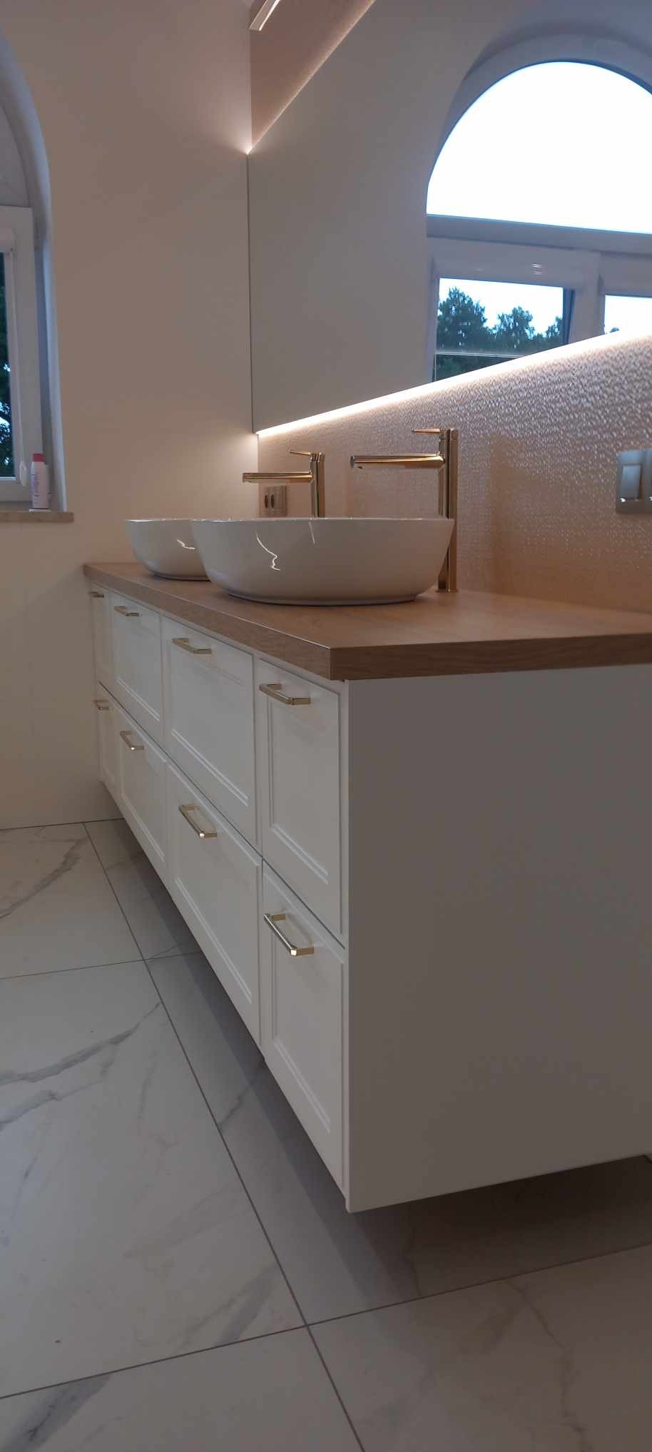 Nowoczesna łazienka z podwójną umywalką nablatową, białe szafki z frezowanymi frontami i złotymi uchwytami, drewniany blat, oświetlenie LED nad lustrem i marmurowe płytki podłogowe.