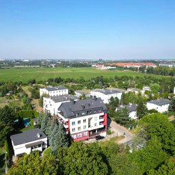 Poter Nieruchomości sp. z o.o - Widok z lotu ptaka na nowoczesny, trzypiętrowy budynek mieszkalny z czerwonymi akcentami, otoczony zielenią drzew i pól, pod błękitnym niebem. Osiedle domów w tle.