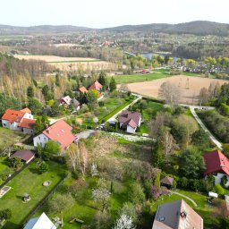 Poter Nieruchomości sp. z o.o - Panoramiczny widok z lotu ptaka na malowniczą wieś z domami z czerwonymi dachami, otoczoną zielenią drzew i pól, w tle jezioro i wzgórza. Słoneczny dzień.