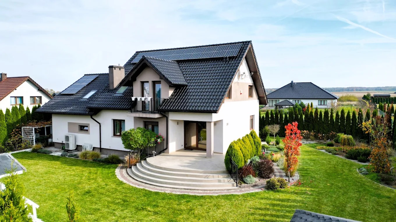 Nowoczesny dom jednorodzinny z panelami słonecznymi na dachu, otoczony zadbanym ogrodem z tujami i kolorowymi krzewami. Widok z góry na taras i schody.