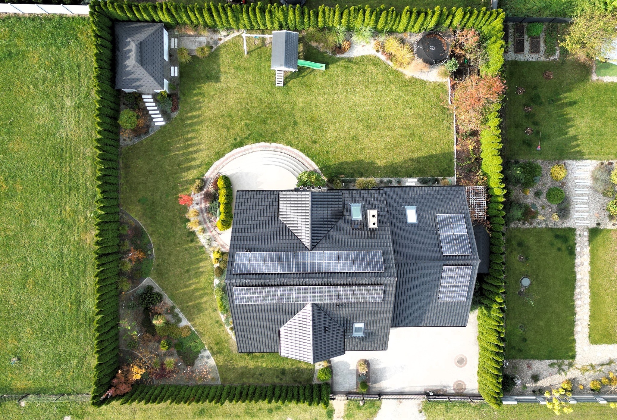 Zdjęcie z drona przedstawia dom z ciemnym dachem i panelami słonecznymi, otoczony zielonym trawnikiem i żywopłotem. Widoczny plac zabaw i domek narzędziowy.