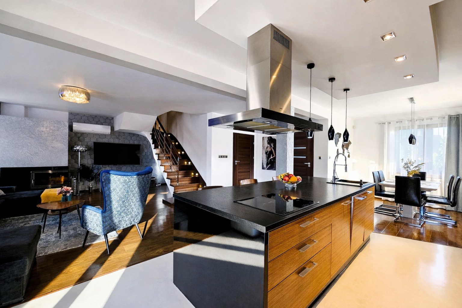 Przestronne wnętrze domu z otwartą kuchnią, wyspą kuchenną z czarnym blatem i drewnianymi szafkami, schodami z drewnianymi stopniami i salonem z kominkiem.