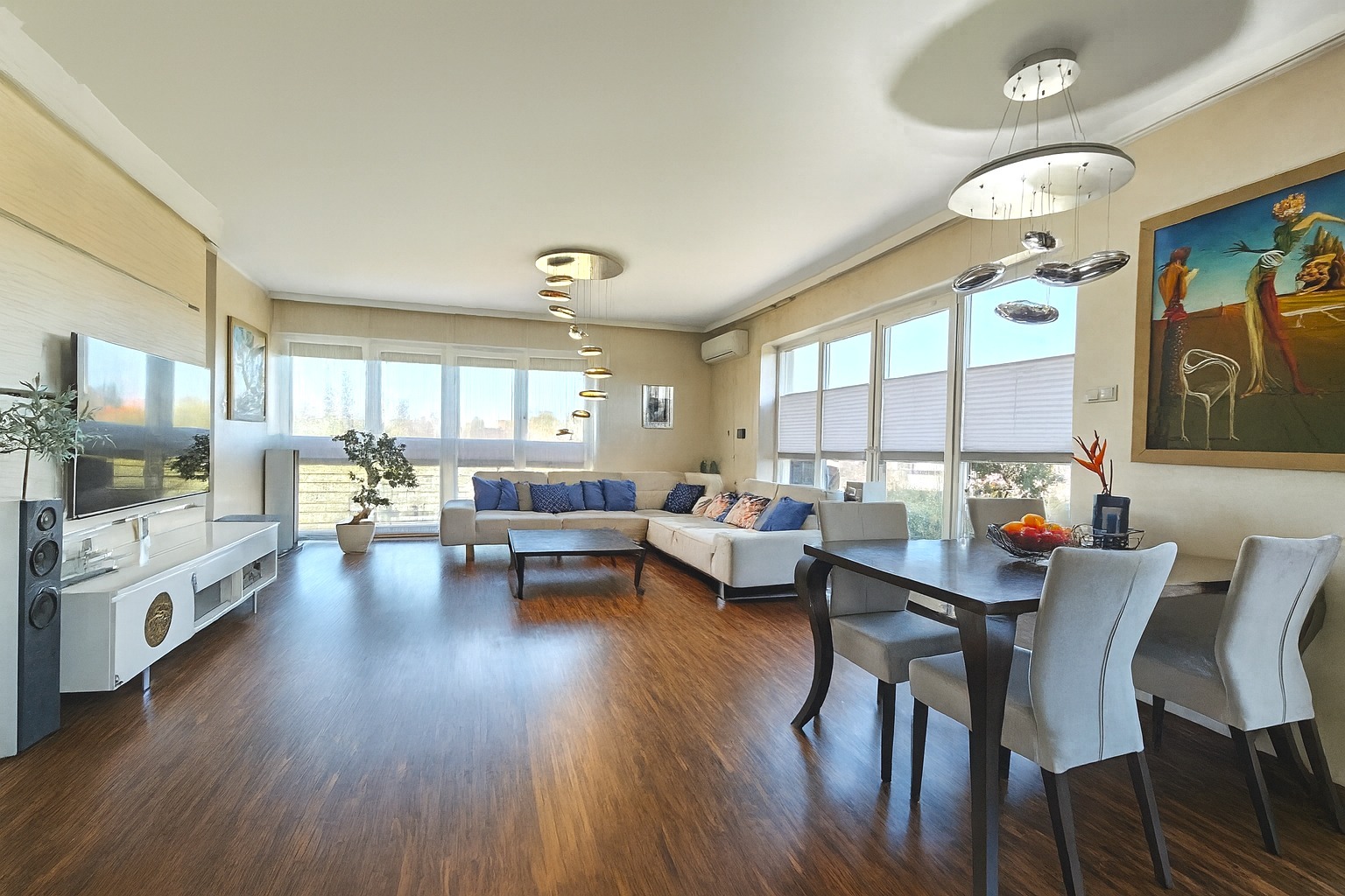 Jasne, przestronne wnętrze salonu z drewnianą podłogą, narożną sofą z niebieskimi poduszkami i stołem jadalnym. Duże okna z roletami wpuszczają dużo światła.