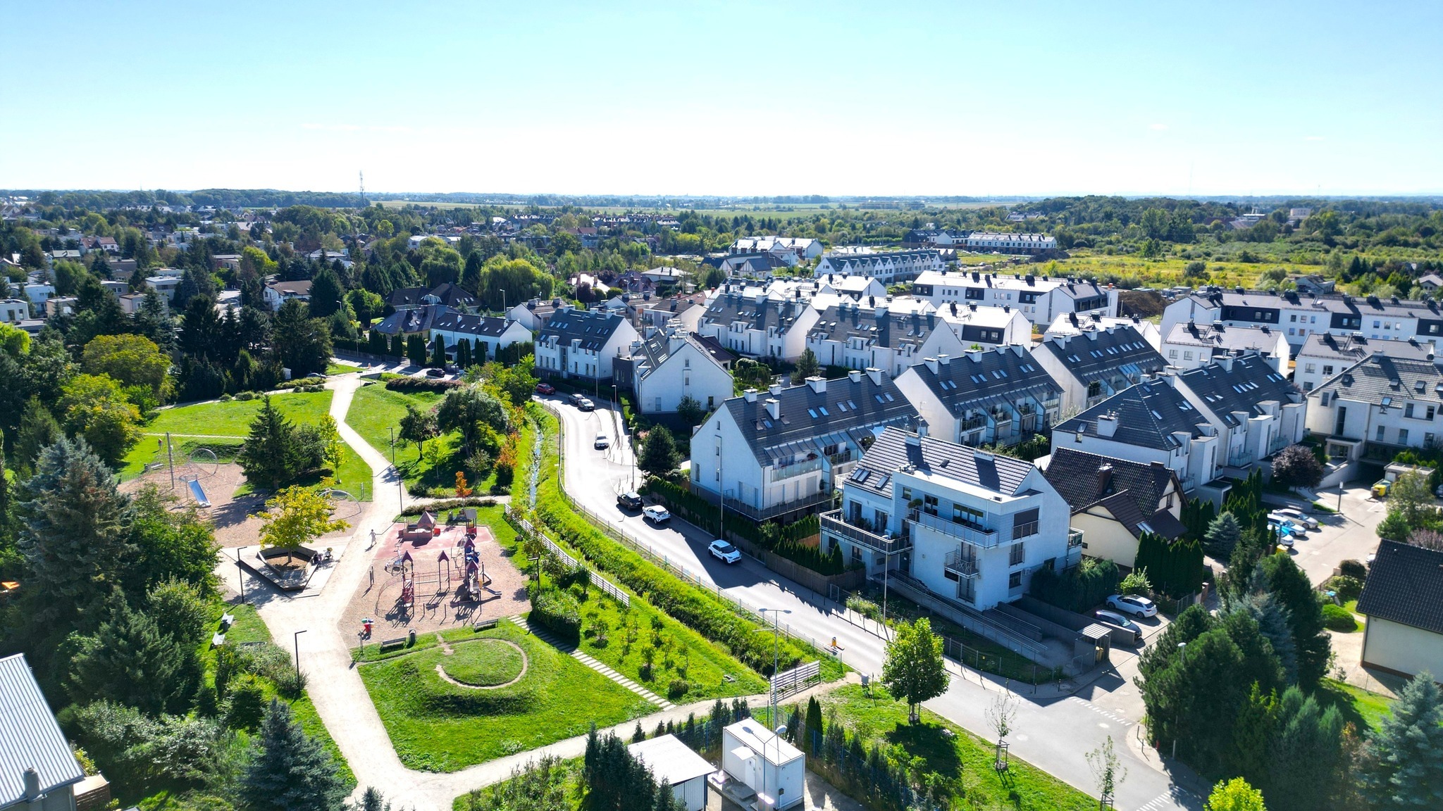 Osiedle nowoczesnych, białych domów z ciemnymi dachami, widziane z lotu ptaka. W tle zieleń drzew i błękitne niebo. Uwagę przykuwa plac zabaw po lewej stronie.