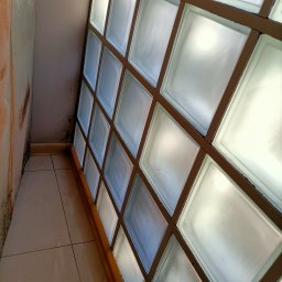 Martyna Kołucka Operacja Błysk - Ściana z luksferami z brązową ramą, widoczne zabrudzenia na ścianie obok i płytkach podłogowych.