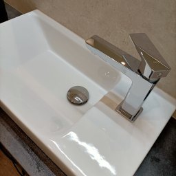 Martyna Kołucka Operacja Błysk - Biała, prostokątna umywalka z chromowaną baterią o kwadratowej podstawie i korkiem, na blacie o fakturze kamienia.