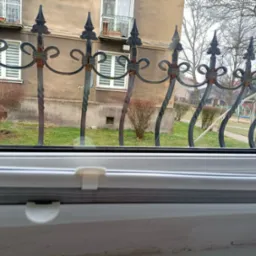Widok z okna na ozdobny, metalowy płot z elementami rdzy, budynek mieszkalny i drzewa w tle.