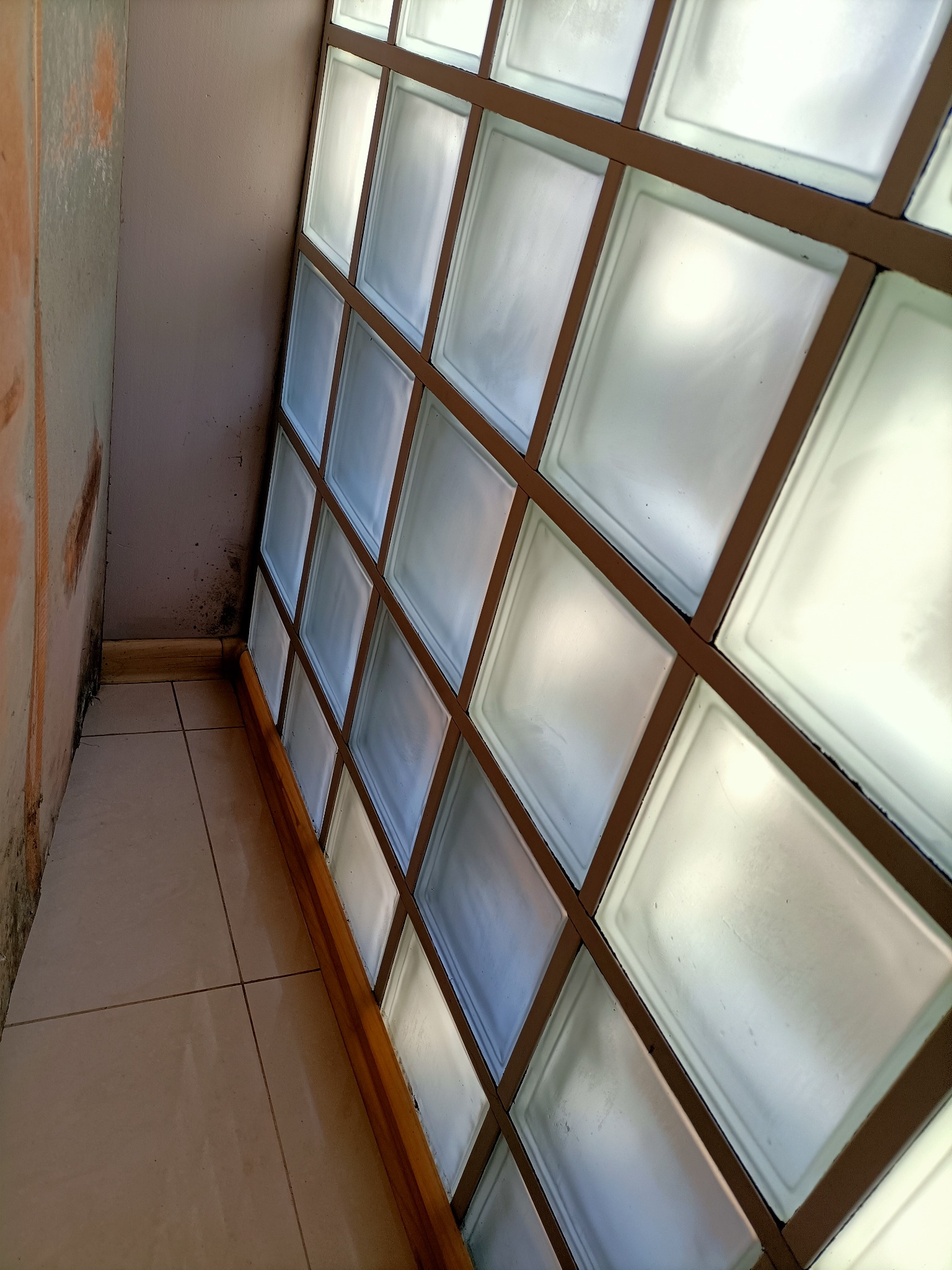 Ściana z luksferami z brązową ramą, widoczne zabrudzenia na ścianie obok i płytkach podłogowych.