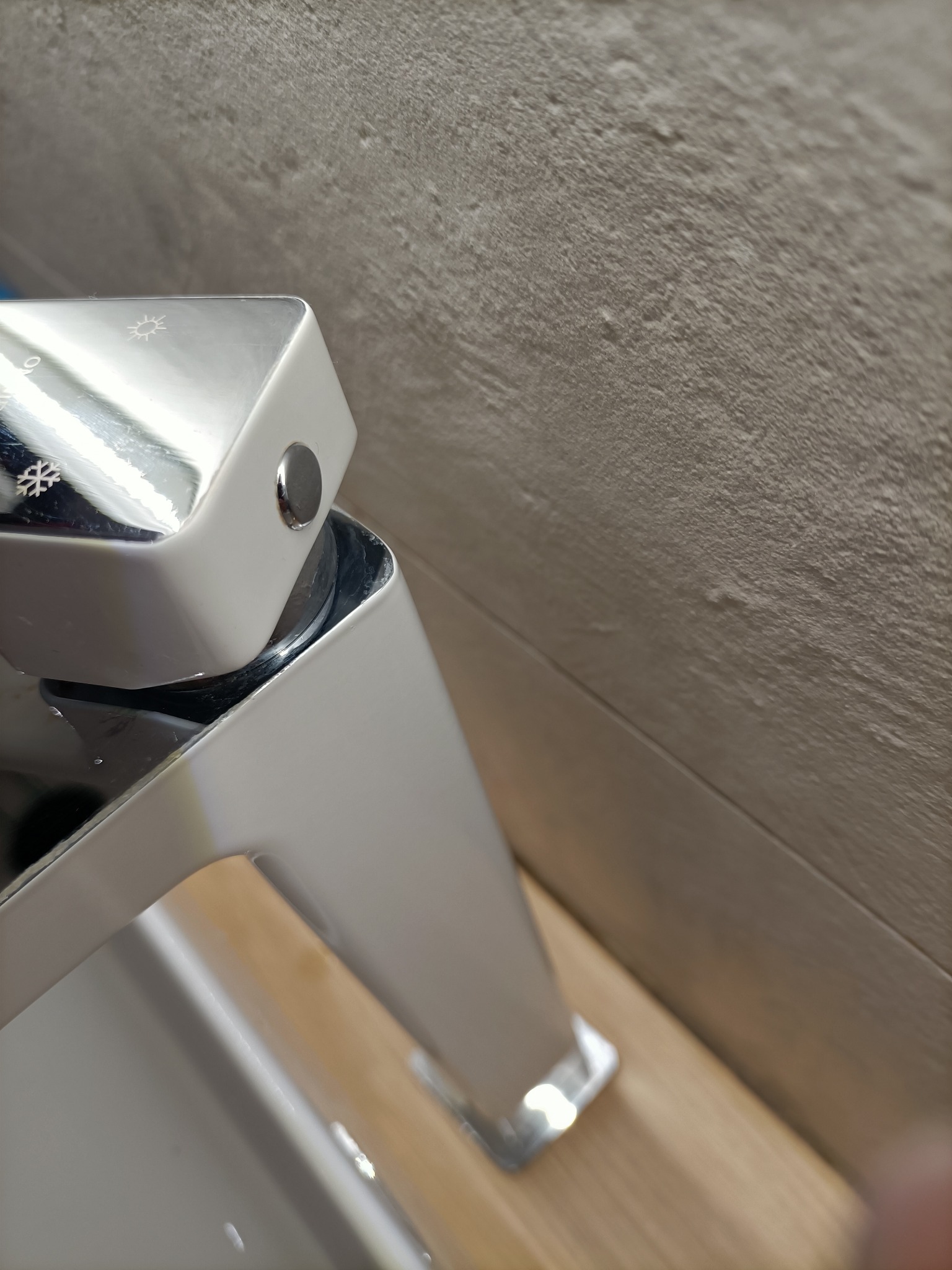 Błyszcząca, chromowana bateria umywalkowa z regulacją temperatury (słońce/śnieżynka) na tle beżowej ściany wykończonej płytkami imitującymi kamień.