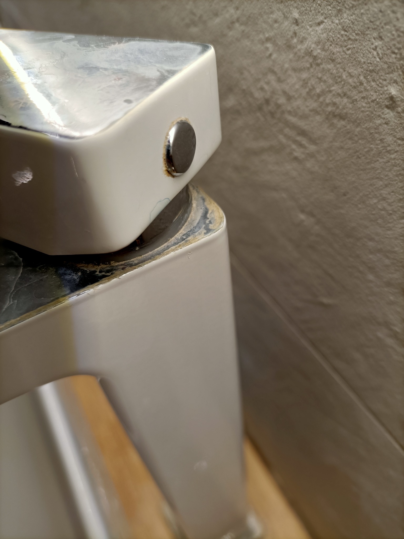 Niedoczyszczona bateria umywalkowa z widocznymi osadami kamienia i zabrudzeniami przy podstawie, na tle szarej ściany.