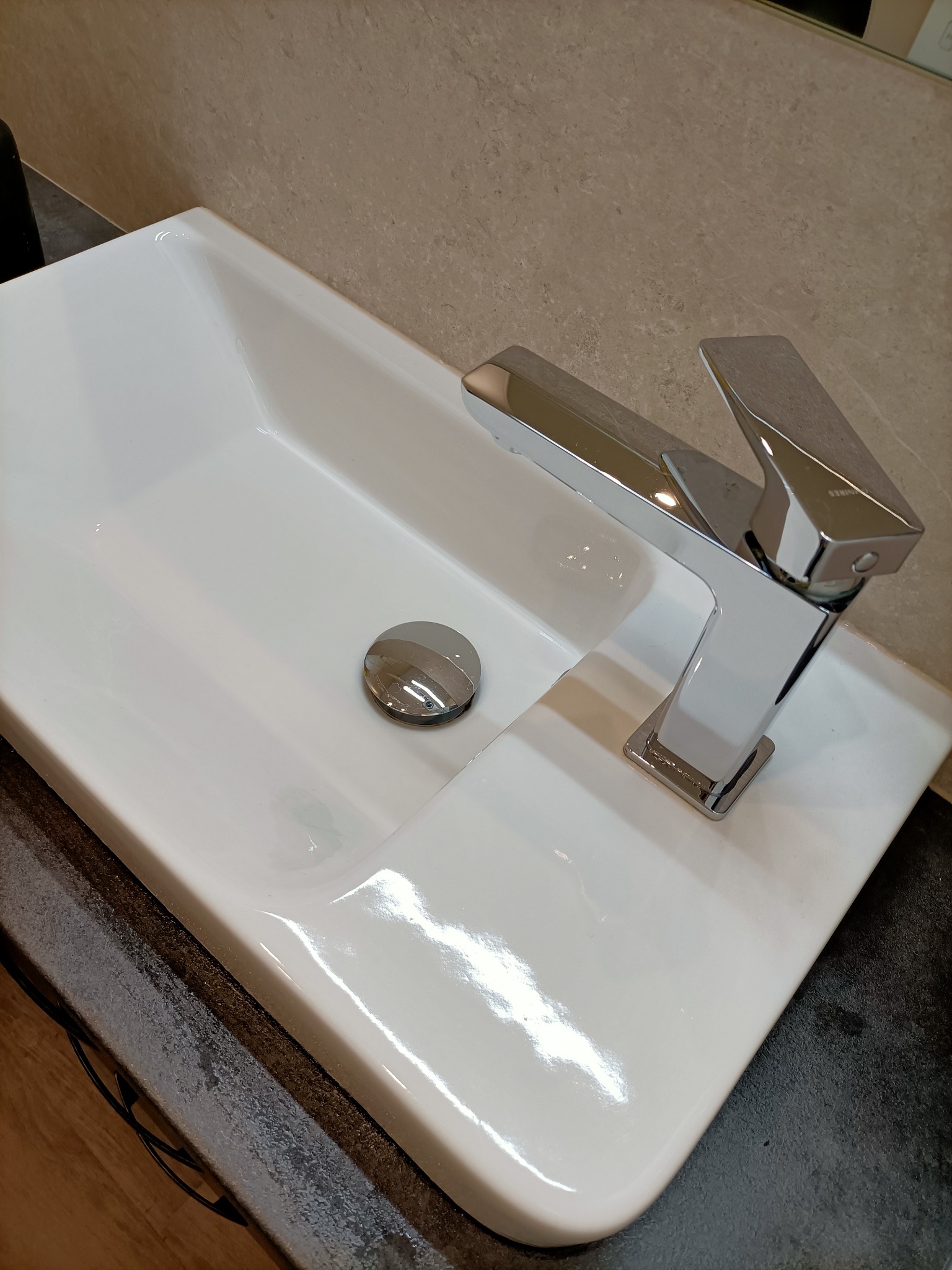 Biała, prostokątna umywalka z chromowaną baterią o kwadratowej podstawie i korkiem, na blacie o fakturze kamienia.