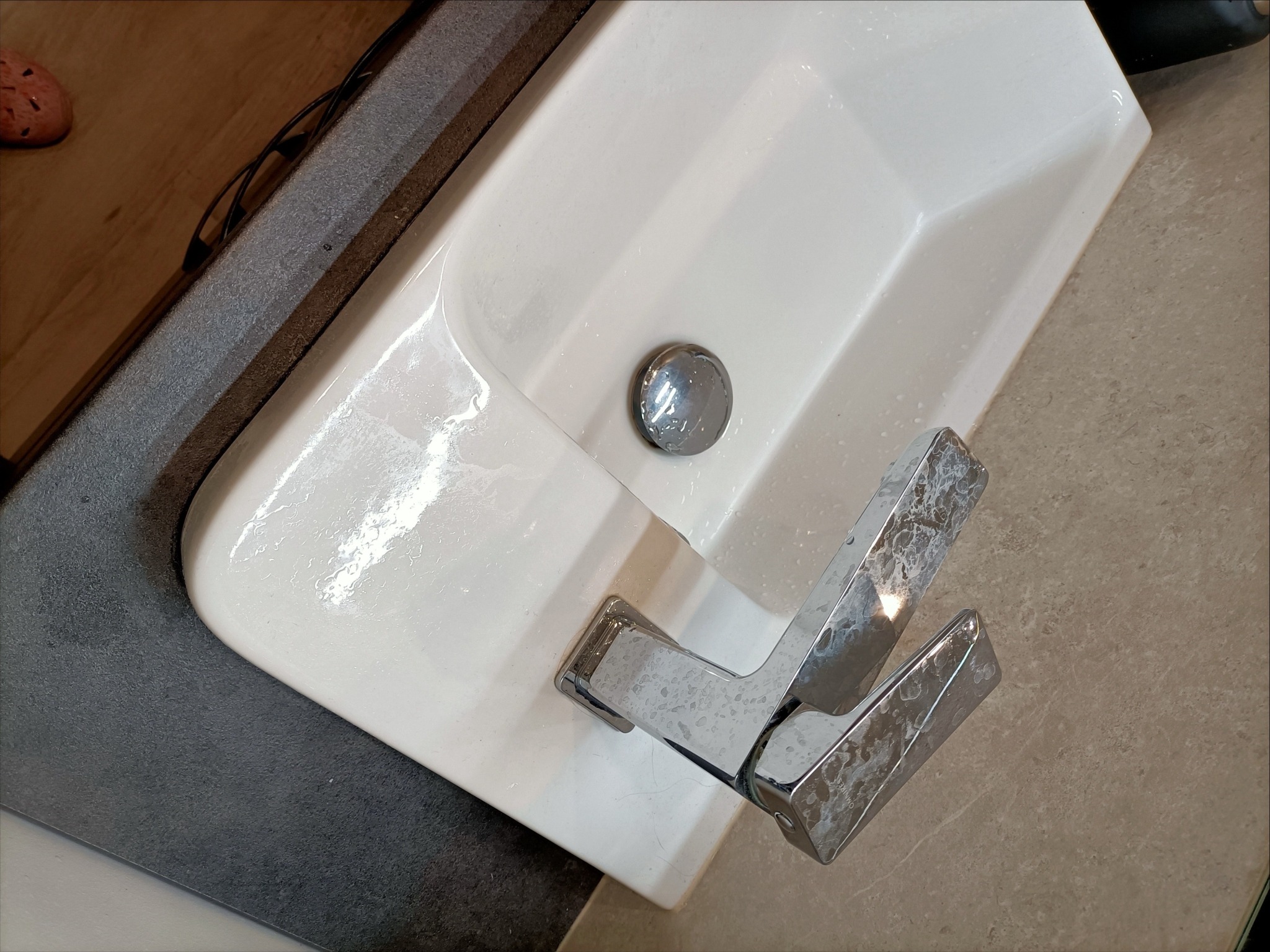 Biała umywalka z chromowaną baterią, na której widoczne są ślady zaschniętej wody, stojąca na blacie z szarego kamienia.