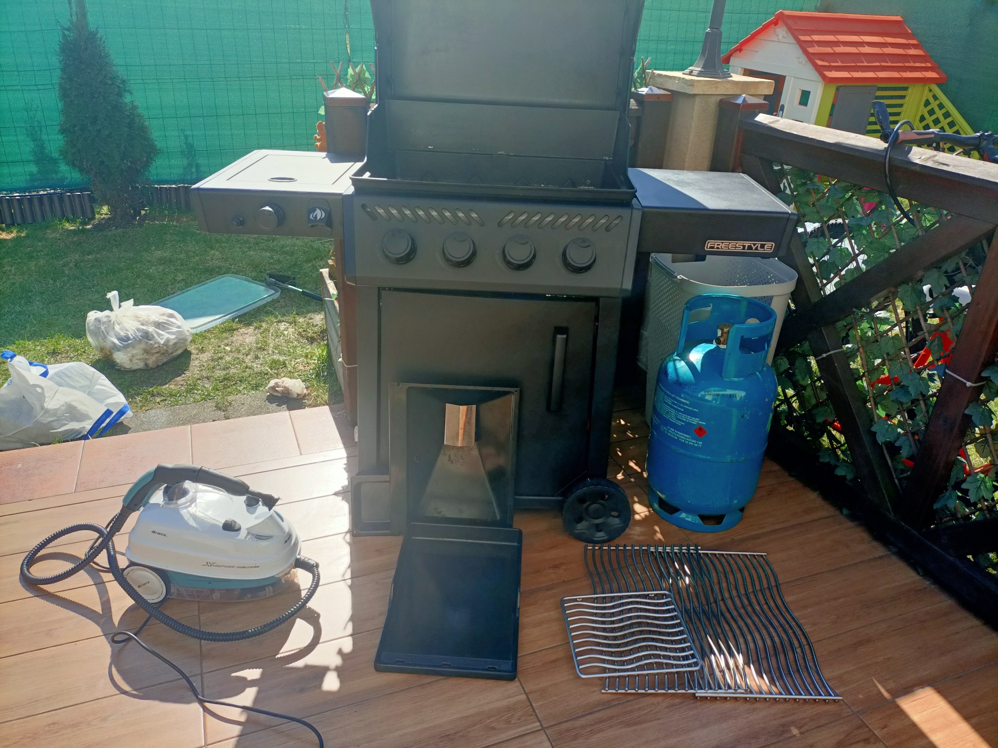 Czyszczenie grilla parownicą: parownica, elementy grilla, butla gazowa i grill na drewnianym tarasie.