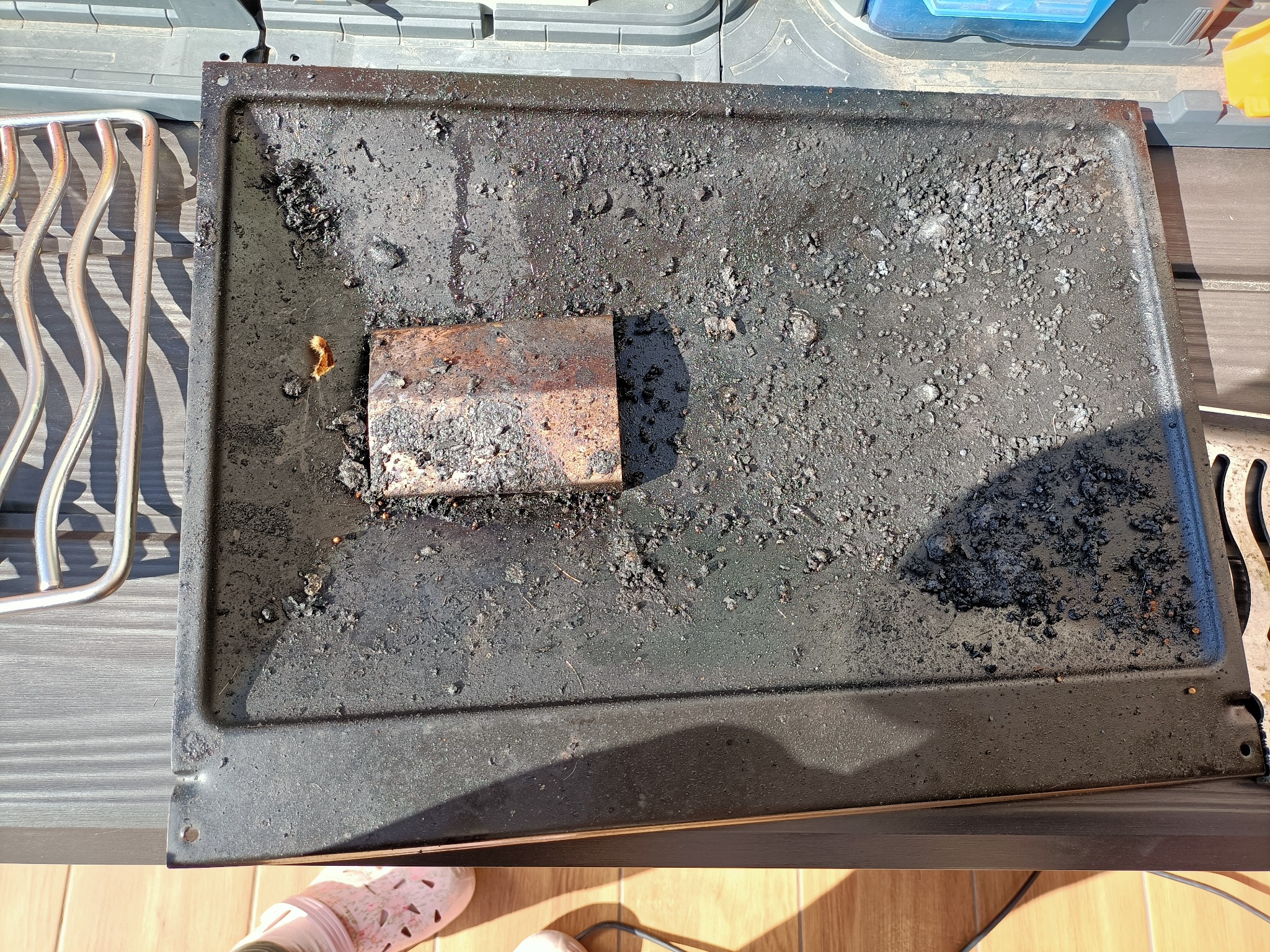 Bardzo zabrudzona, czarna blacha piekarnika z przypalonymi resztkami jedzenia i tłuszczu, obok metalowy ruszt.