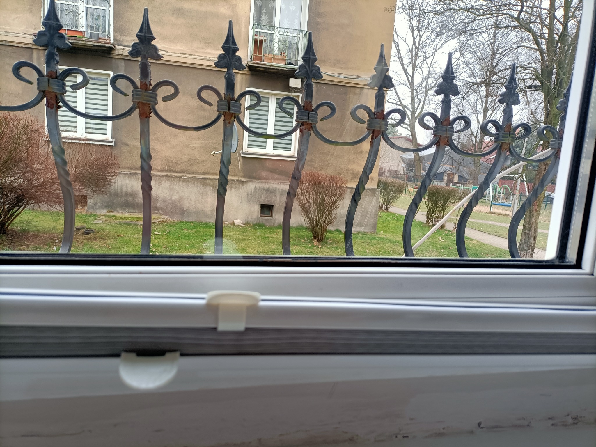 Widok z okna na ozdobny, metalowy płot z elementami rdzy, budynek mieszkalny i drzewa w tle.