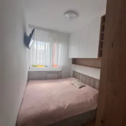 Sypialnia z zabudową meblową: białe szafki wiszące, drewniana półka i regał. Na ścianie telewizor, obok okno z firankami, łóżko z różową narzutą.