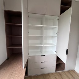 MD STUDIO MEBLE NA WYMIAR Maja Dziedzic - Jasna szafa wnękowa z otwartymi drzwiami, widoczne półki i szuflady z czarnymi uchwytami. Obok biurko z drewnianym blatem. Podłoga z paneli.