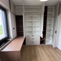 MD STUDIO MEBLE NA WYMIAR Maja Dziedzic - Jasna szafa wnękowa z biurkiem pod oknem, otwarte białe fronty, brązowe półki, drewniana podłoga, nowoczesne oświetlenie sufitowe. Funkcjonalne rozwiązanie do przechowywania.