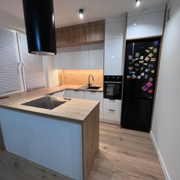 MD STUDIO MEBLE NA WYMIAR Maja Dziedzic - Nowoczesna kuchnia z wyspą i czarnym okapem cylindrycznym. Białe fronty szafek kontrastują z drewnianym blatem i panelami. Czarna lodówka z magnesami.