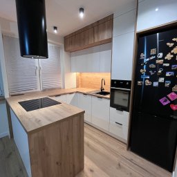 MD STUDIO MEBLE NA WYMIAR Maja Dziedzic - Nowoczesna kuchnia z wyspą: białe szafki, drewniane blaty, czarny okap cylindryczny i lodówka z magnesami. Minimalistyczny design, ciepłe oświetlenie.