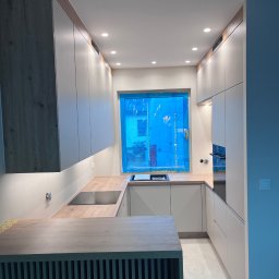 MD STUDIO MEBLE NA WYMIAR Maja Dziedzic - Nowoczesna kuchnia w odcieniach bieli i drewna z wyspą. Okno zabezpieczone folią ochronną. Oświetlenie punktowe podkreśla minimalistyczny design.