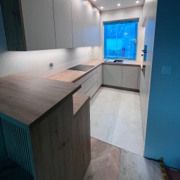 MD STUDIO MEBLE NA WYMIAR Maja Dziedzic - Jasna kuchnia z drewnianym blatem i wyspą. Okno zabezpieczone folią. Nowoczesne oświetlenie podszafkowe. Parkiet ułożony w jodełkę. Minimalistyczny design.