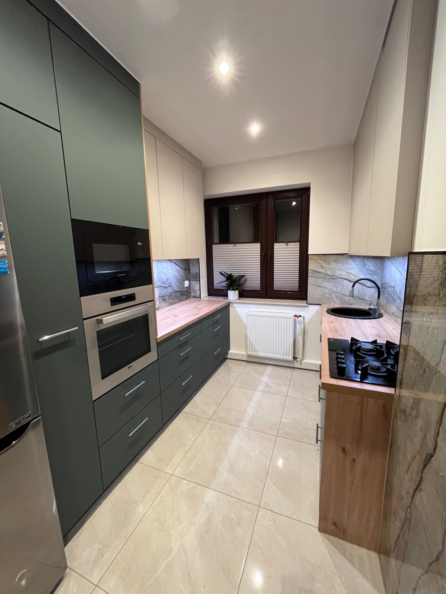 Nowoczesna kuchnia z zielonymi i beżowymi szafkami, drewnianym blatem i płytą gazową. Widoczne okno z żaluzjami plisowanymi i marmurowe wykończenie ściany.