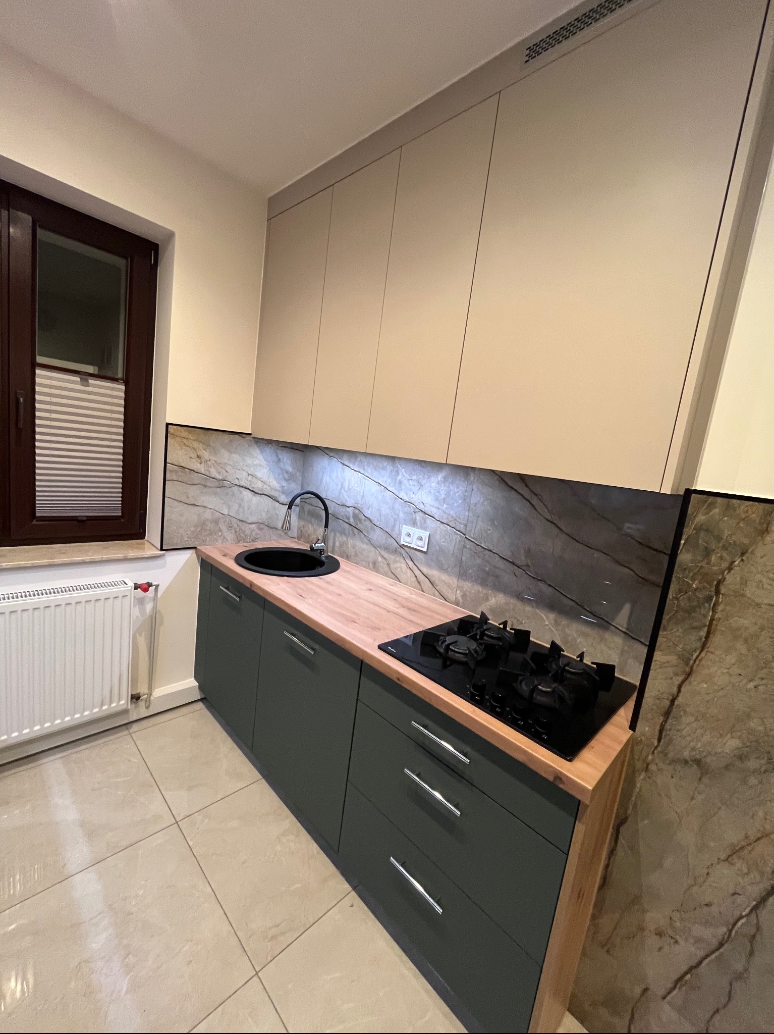 Nowoczesna kuchnia z ciemnozielonymi szafkami, drewnianym blatem i marmurowym panelem nad blatem. Widoczny zlew, płyta grzewcza i okno z żaluzjami.