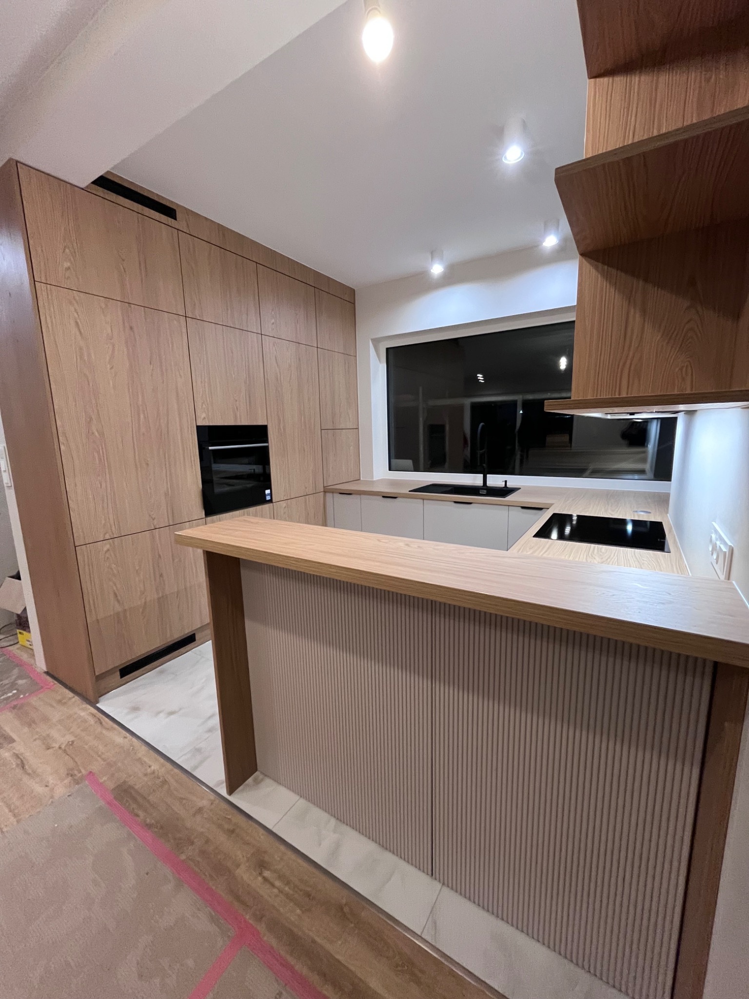 Jasna kuchnia z drewnianymi frontami i wyspą z ryflowanym wykończeniem. Widoczne sprzęty AGD i duże okno. Podłoga w trakcie wykończenia.