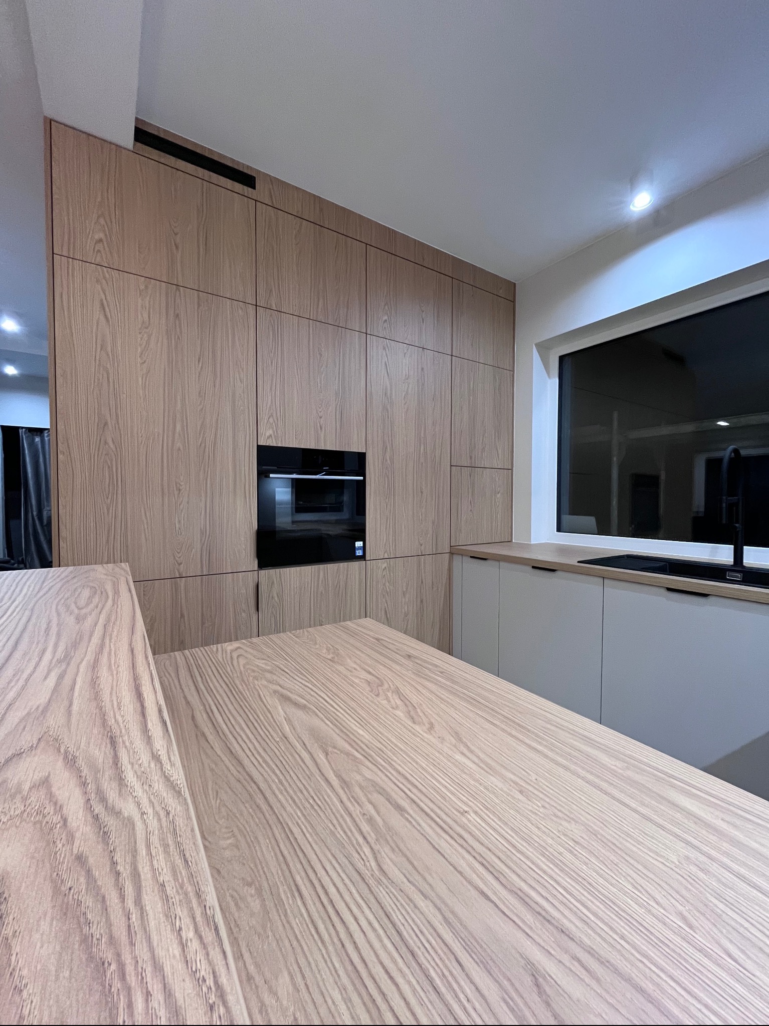 Nowoczesna kuchnia z drewnianymi frontami, wbudowanym piekarnikiem i czarną armaturą przy oknie. Blat z wyraźnym rysunkiem drewna. Minimalistyczny design.