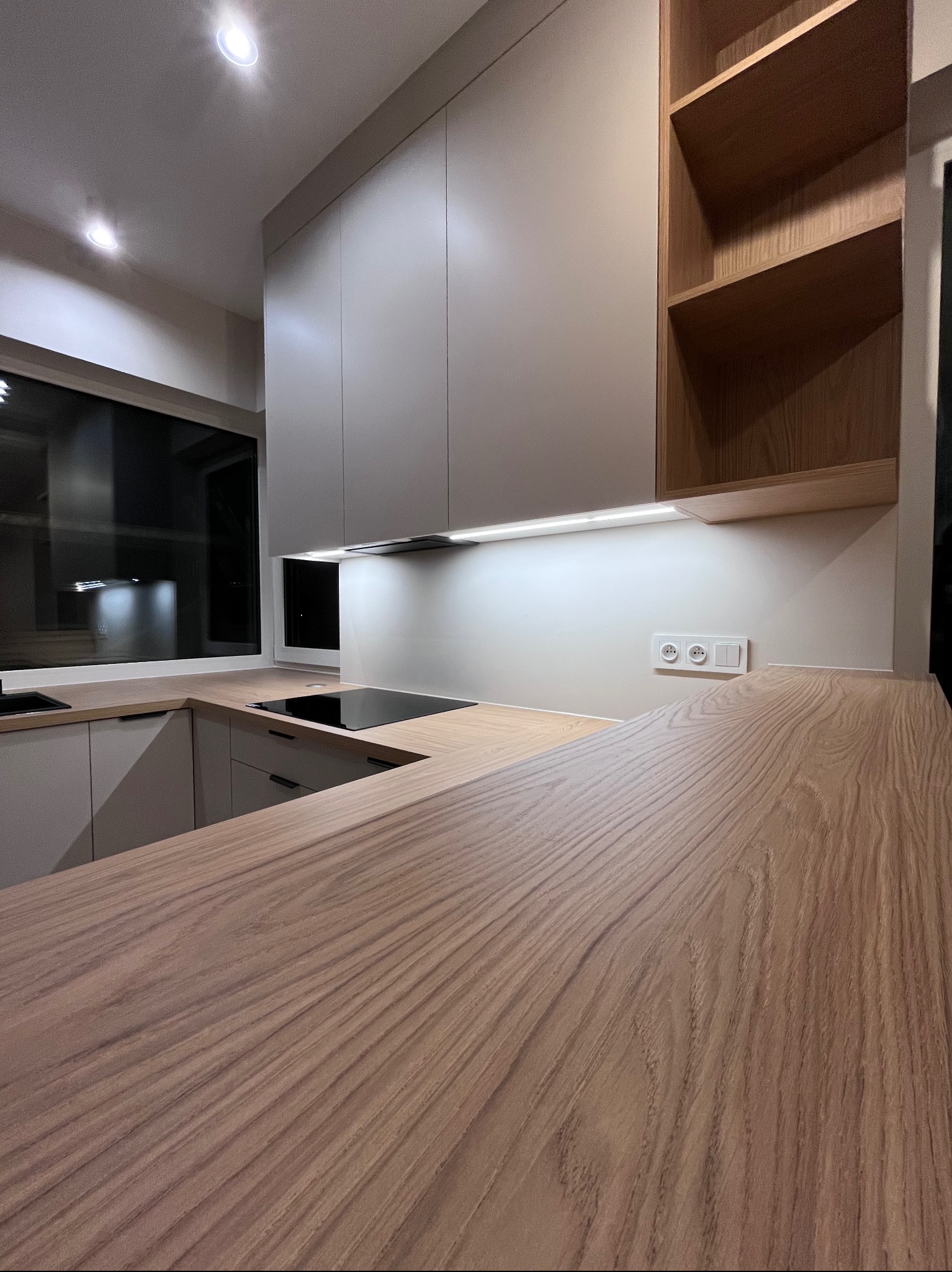 Nowoczesna kuchnia z drewnianym blatem i białymi szafkami, oświetlenie LED pod szafkami, widok na okno i płytę indukcyjną. Minimalistyczny design.