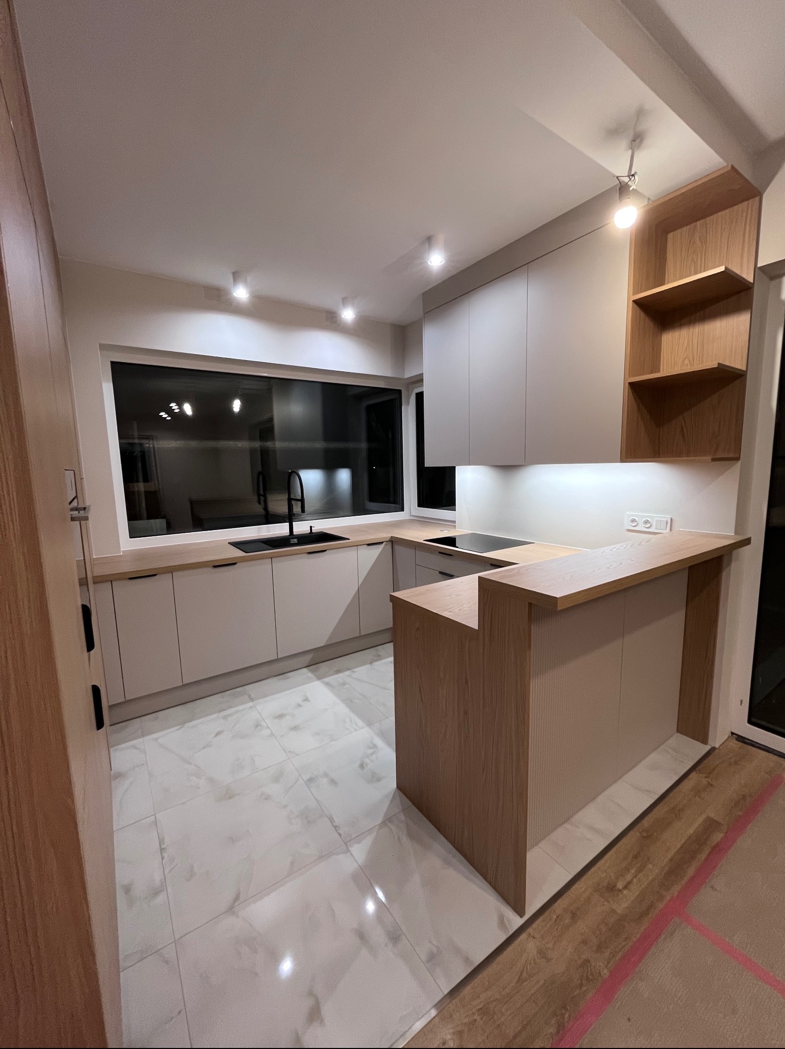 Nowoczesna kuchnia z wyspą i dużym oknem. Jasne fronty szafek, drewniany blat i otwarte półki. Minimalistyczny design, białe płytki podłogowe z marmurkowym wzorem.