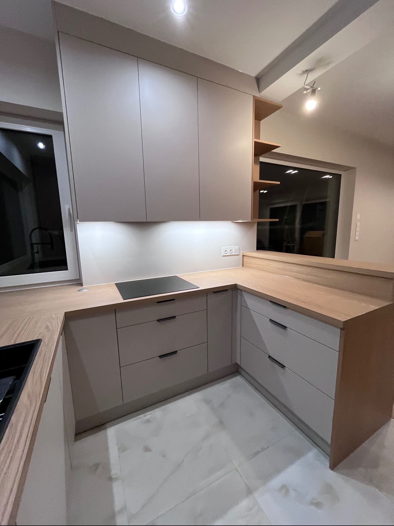 Jasna kuchnia na wymiar z drewnianym blatem i minimalistycznymi uchwytami, oświetlona ledami, z widokiem na okno i marmurową podłogą. Ujęcie z perspektywy.