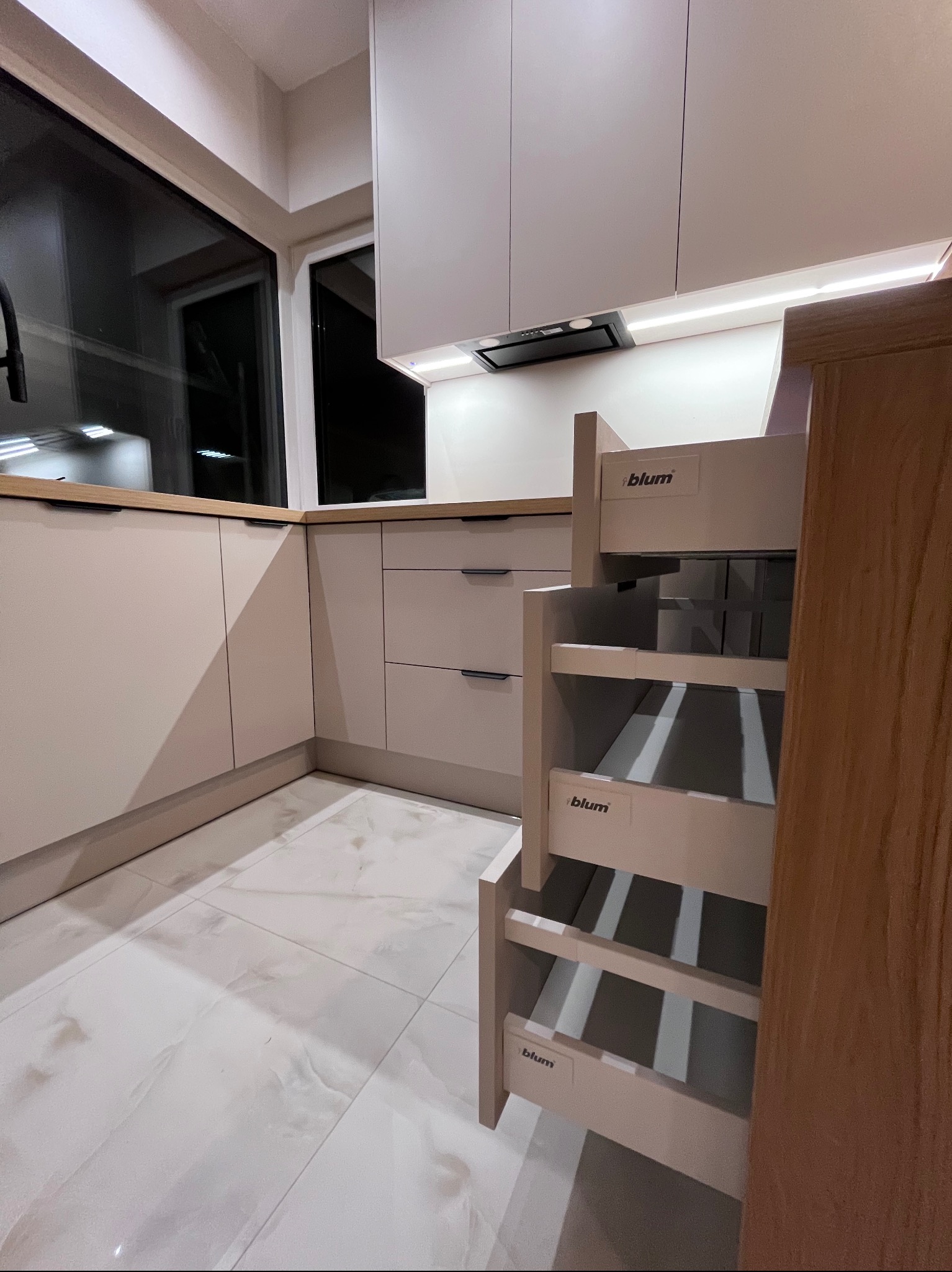 Jasna kuchnia z wysuwanym systemem Blum, widok na okno i płytki podłogowe. Minimalistyczny design, oświetlenie LED pod szafkami górnymi.