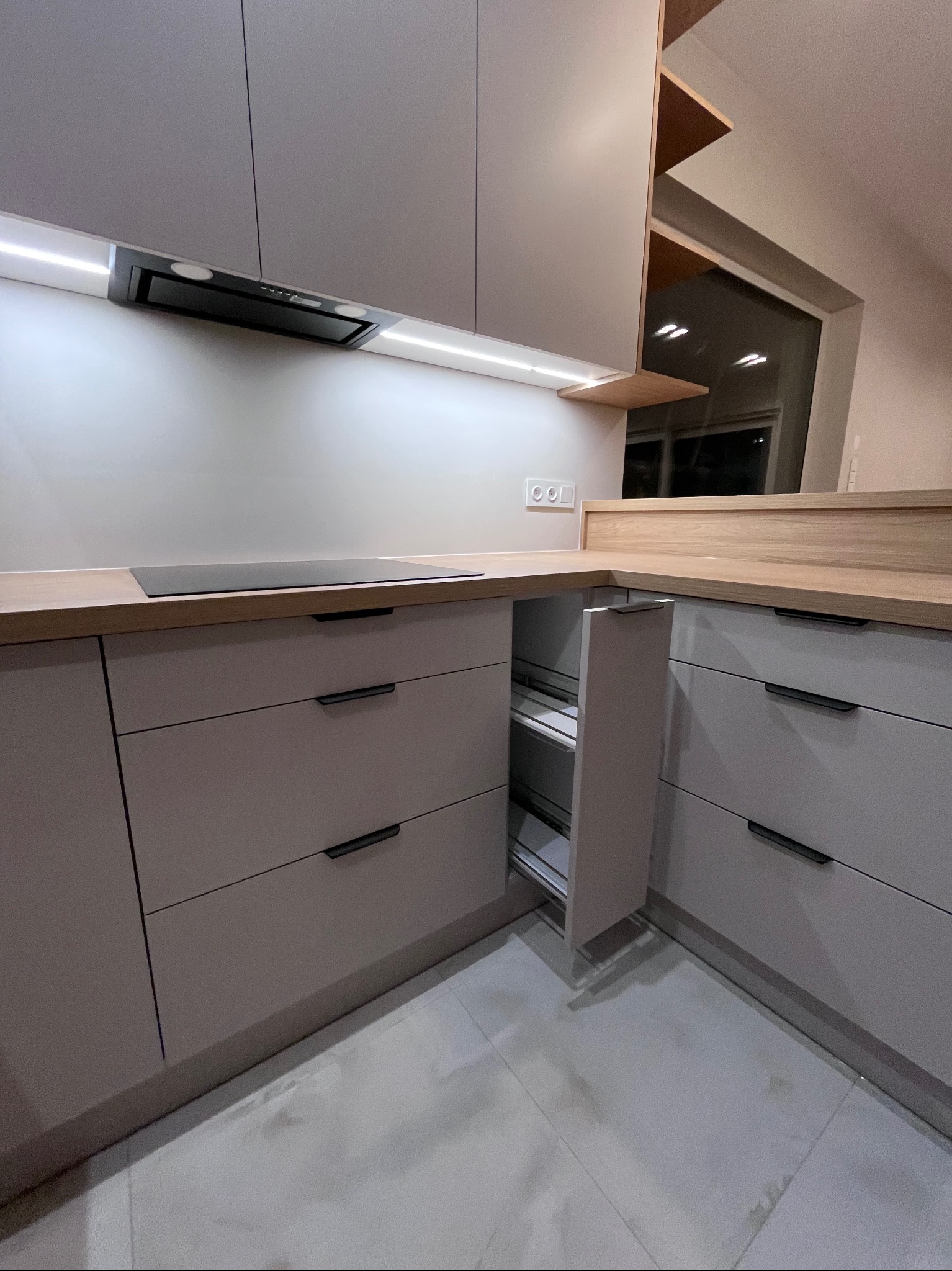 Nowoczesna kuchnia w odcieniach szarości z drewnianym blatem. Wysuwana szafka cargo, płyta indukcyjna i oświetlenie LED pod szafkami wiszącymi. Minimalistyczny design.