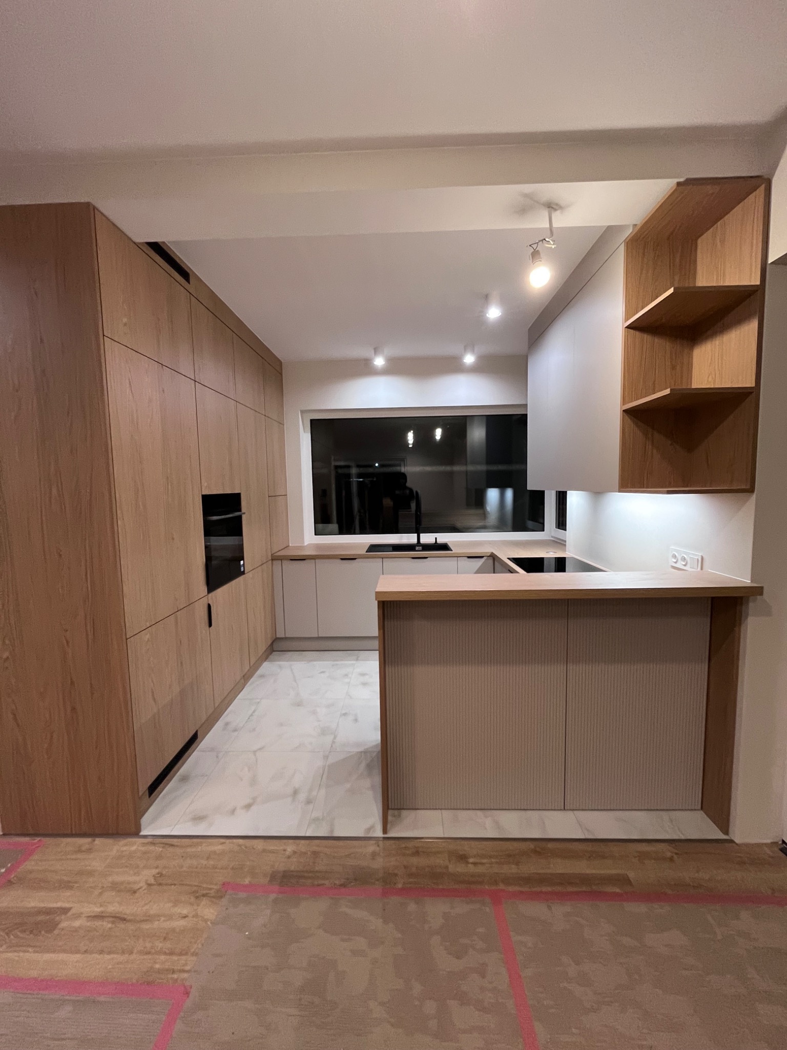 Nowoczesna kuchnia na wymiar z drewnianymi frontami i wyspą, oświetlenie LED, marmurowe płytki podłogowe. Widok na okno i zlew. Minimalistyczny design.