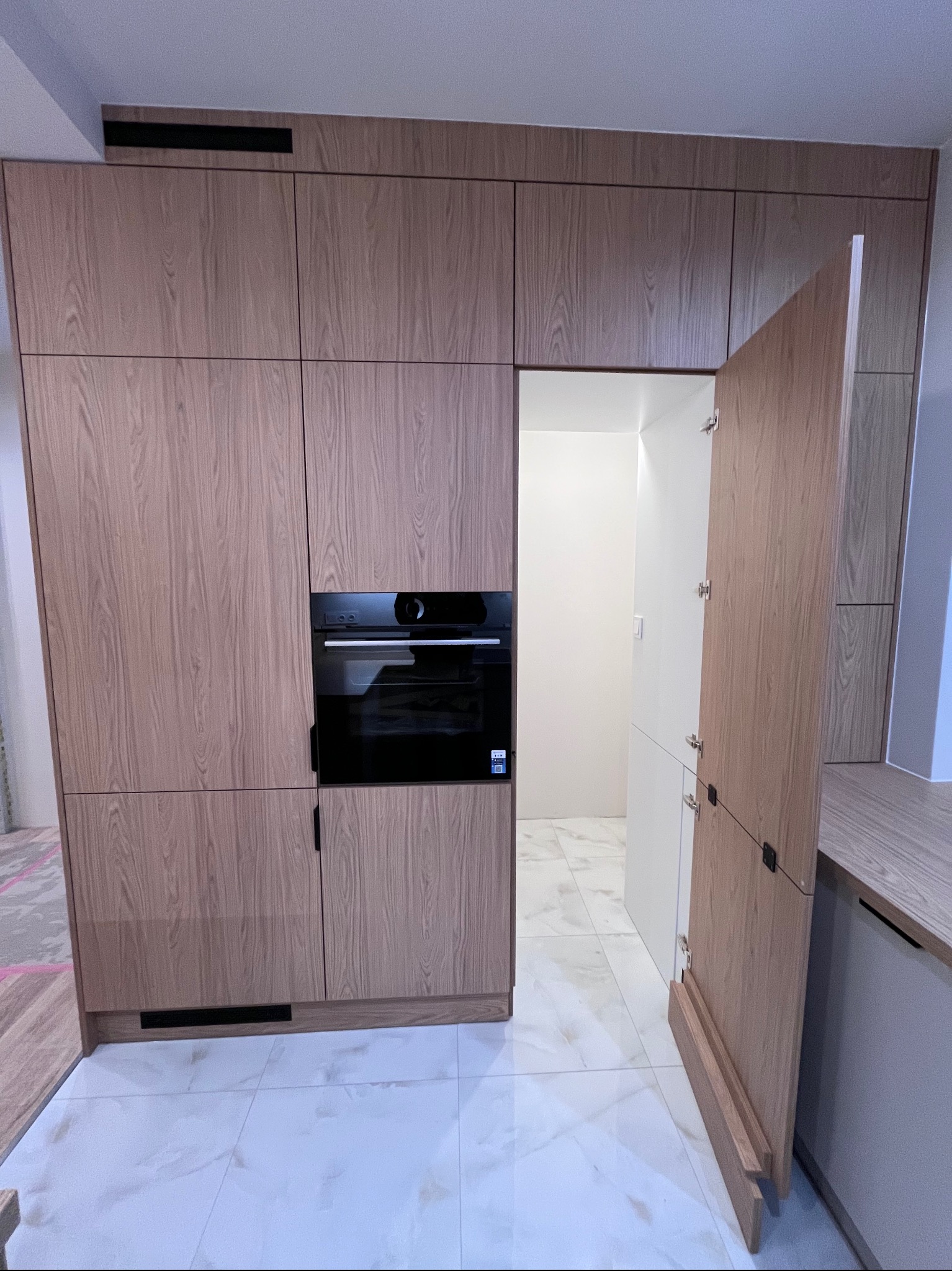 Jasna zabudowa kuchenna z forniru dębowego, z piekarnikiem w zabudowie i ukrytymi drzwiami do pomieszczenia gospodarczego. Biała podłoga z płytek imitujących marmur.