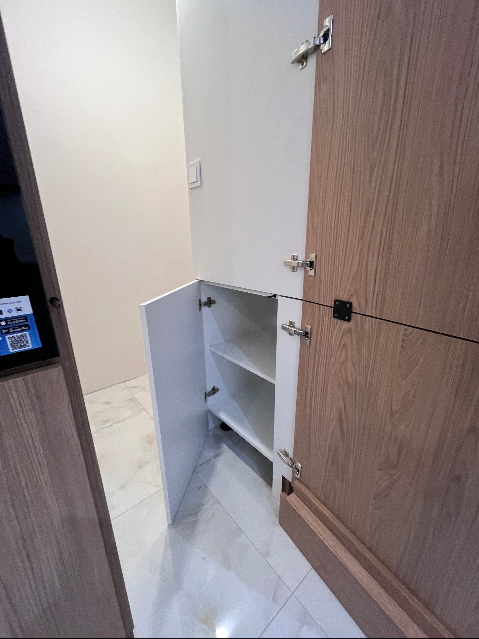 Otwarta szafka z półkami w zabudowie, fronty z drewna i bieli, widoczne okucia. Minimalistyczny design, jasne wnętrze, marmurowe płytki podłogowe.