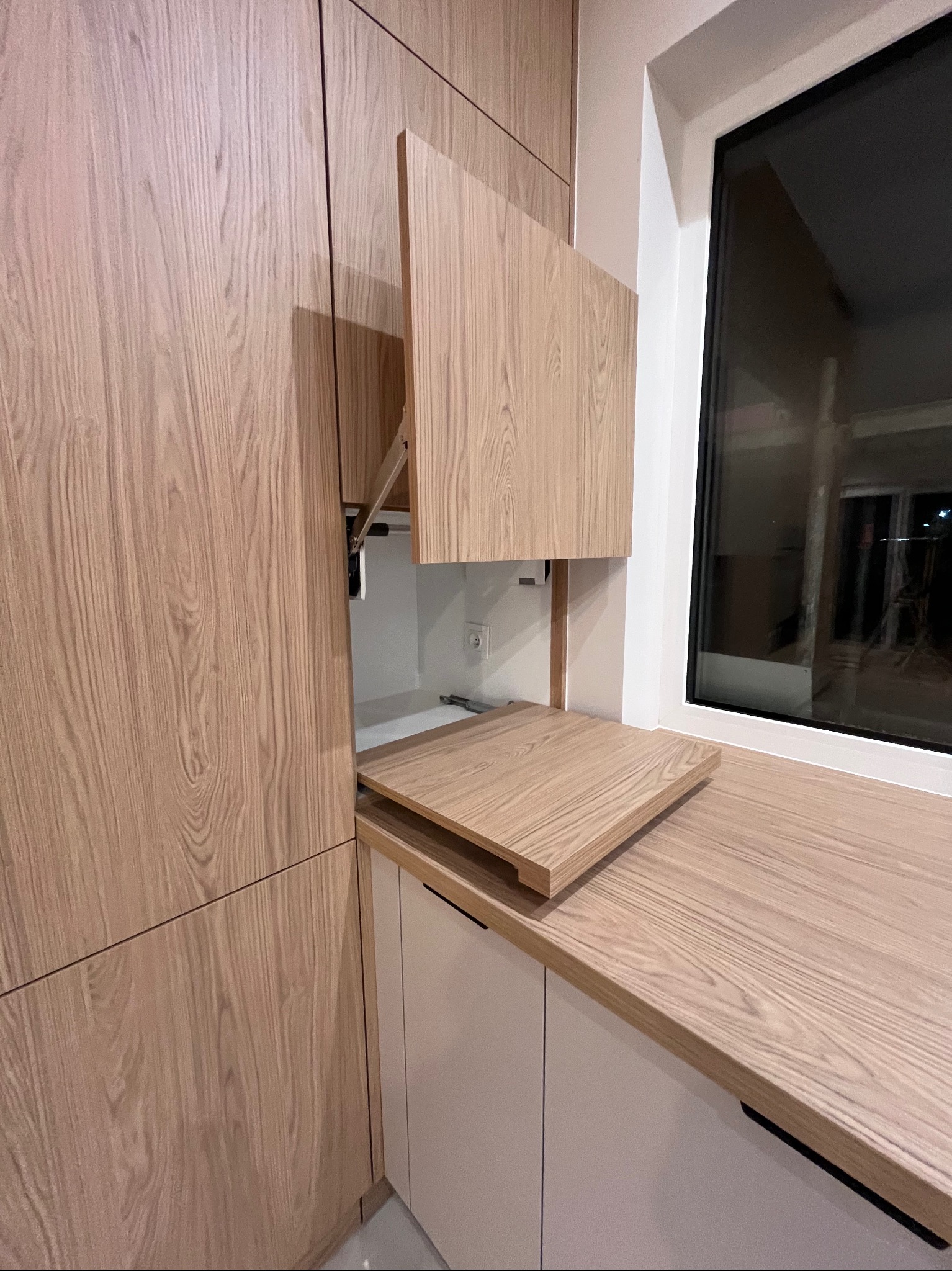 Jasna, drewniana zabudowa kuchenna z otwartą szafką i blatem przy oknie. Widoczne mechanizmy podnoszące szafkę. Minimalistyczny design, ciepłe oświetlenie.