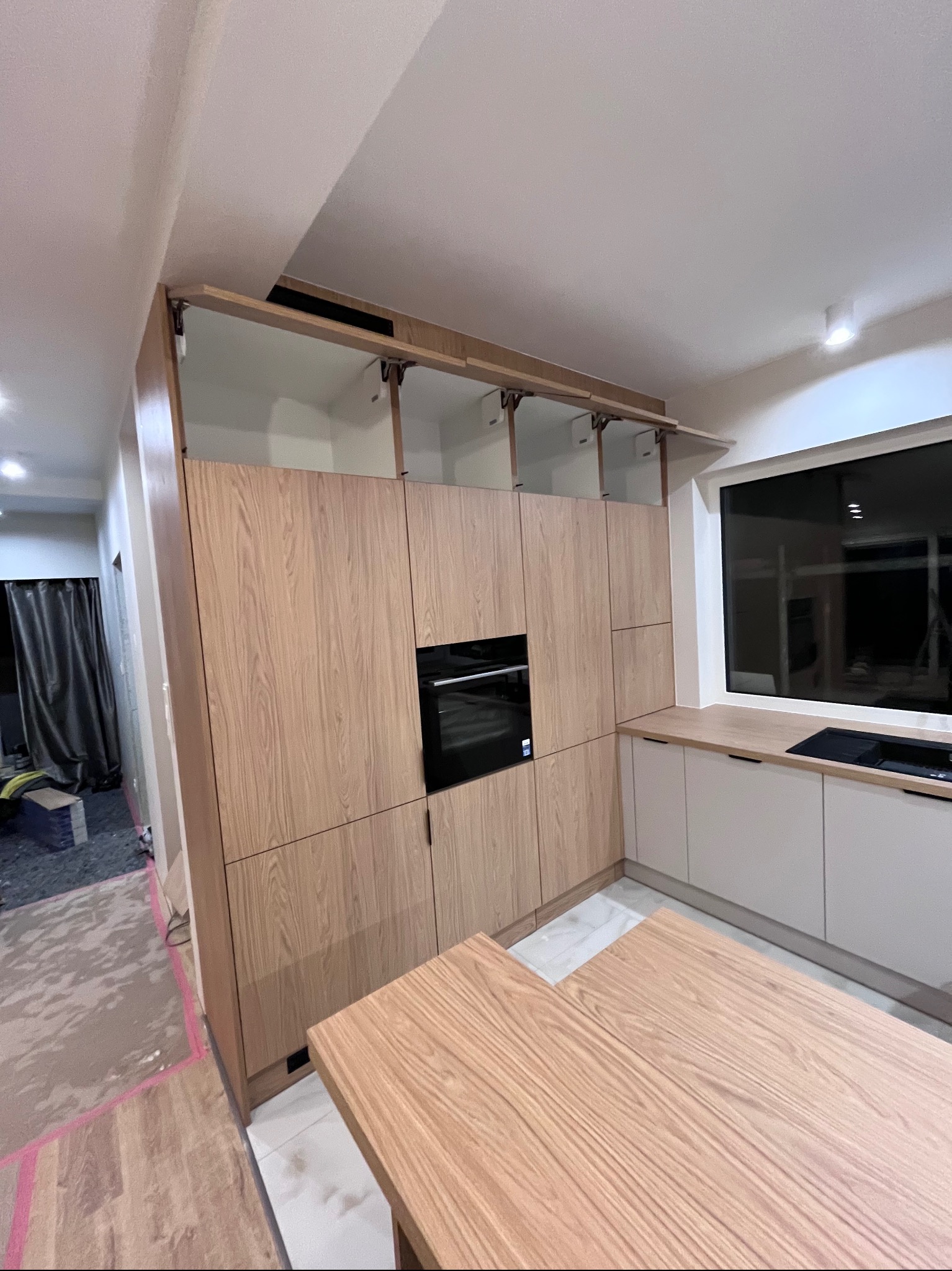 Jasna, drewniana zabudowa kuchenna z otwartymi szafkami górnymi i czarnym piekarnikiem wkomponowanym w szafkę. Widoczny fragment wyspy kuchennej z blatem z tego samego drewna.