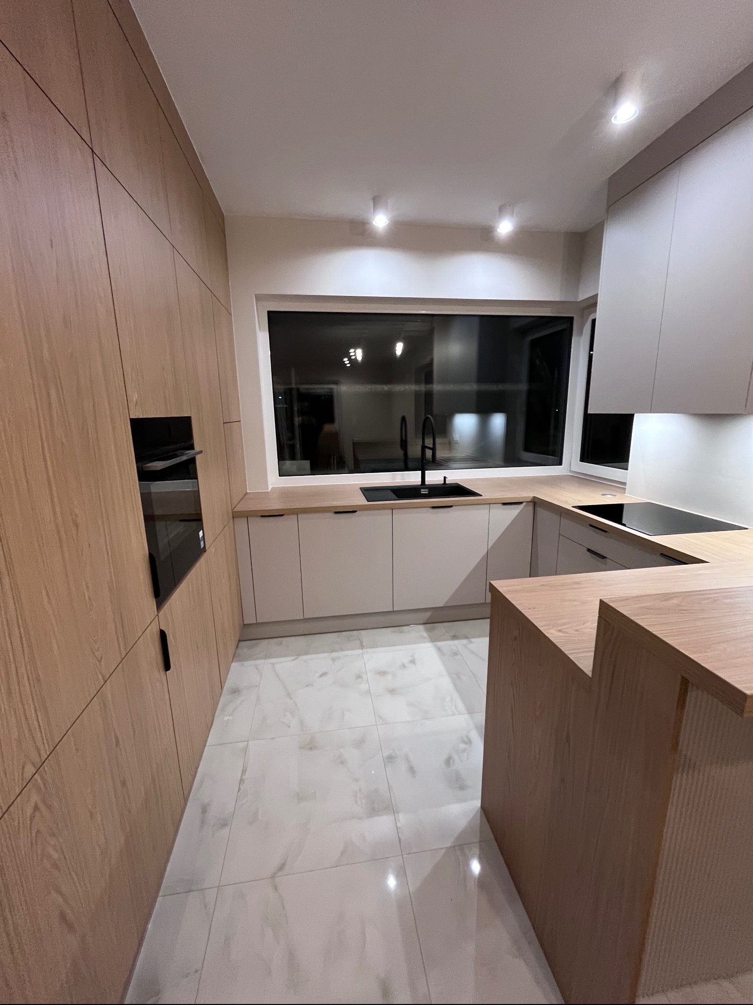 Nowoczesna kuchnia z drewnianymi frontami i wyspą, czarny zlew i płyta indukcyjna, marmurowe płytki podłogowe, minimalistyczne oświetlenie punktowe.