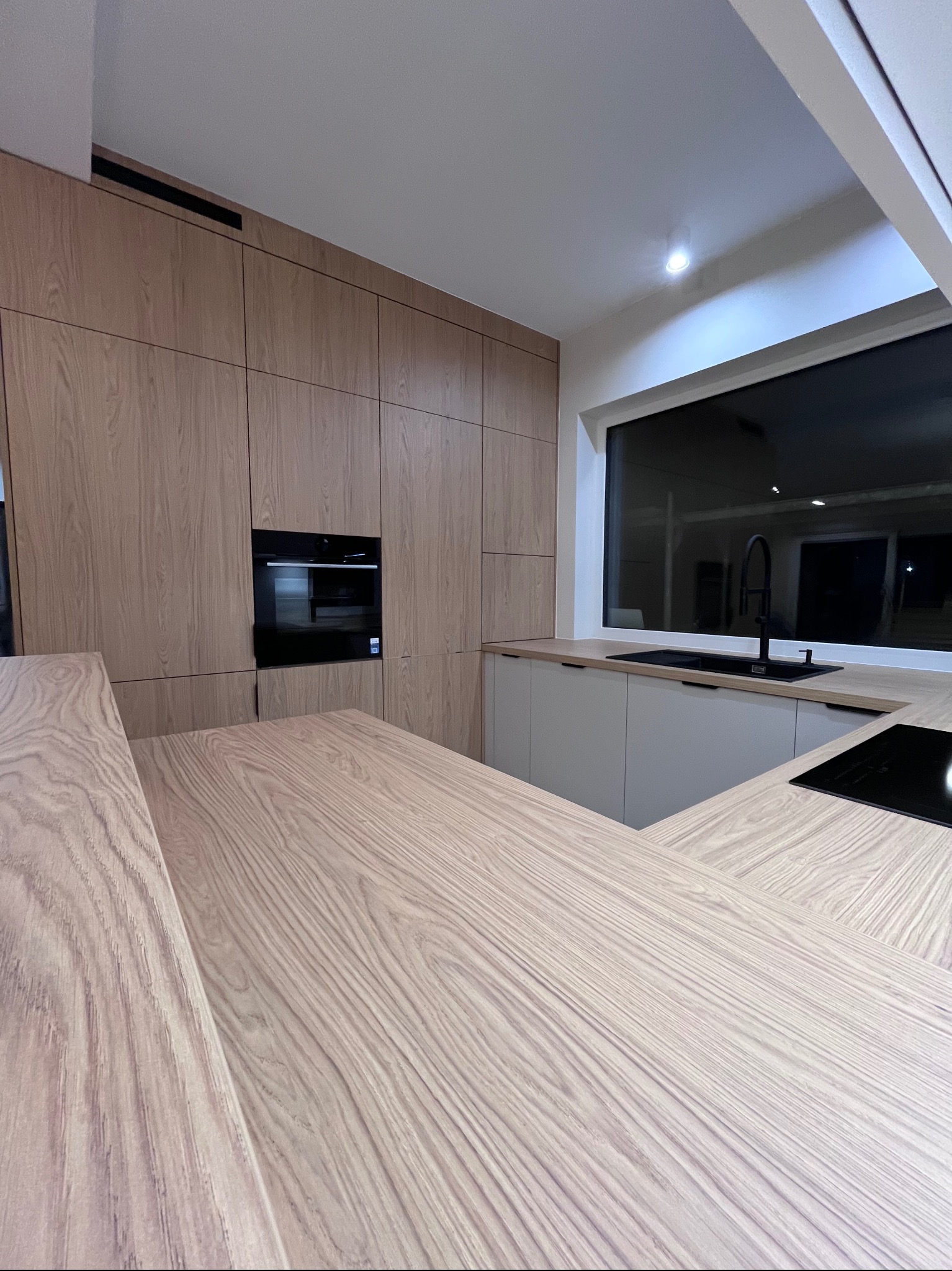 Nowoczesna kuchnia na wymiar z drewnianymi frontami, czarnym piekarnikiem i blatem z wyraźnym usłojeniem. Duże okno z czarną armaturą i płytą grzewczą.